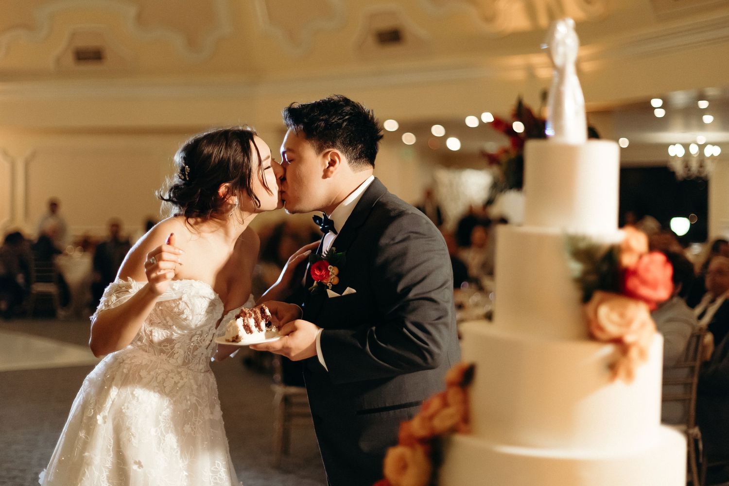 Romantic cake cutting moment in elegant ballroom with tiered white wedding cake.