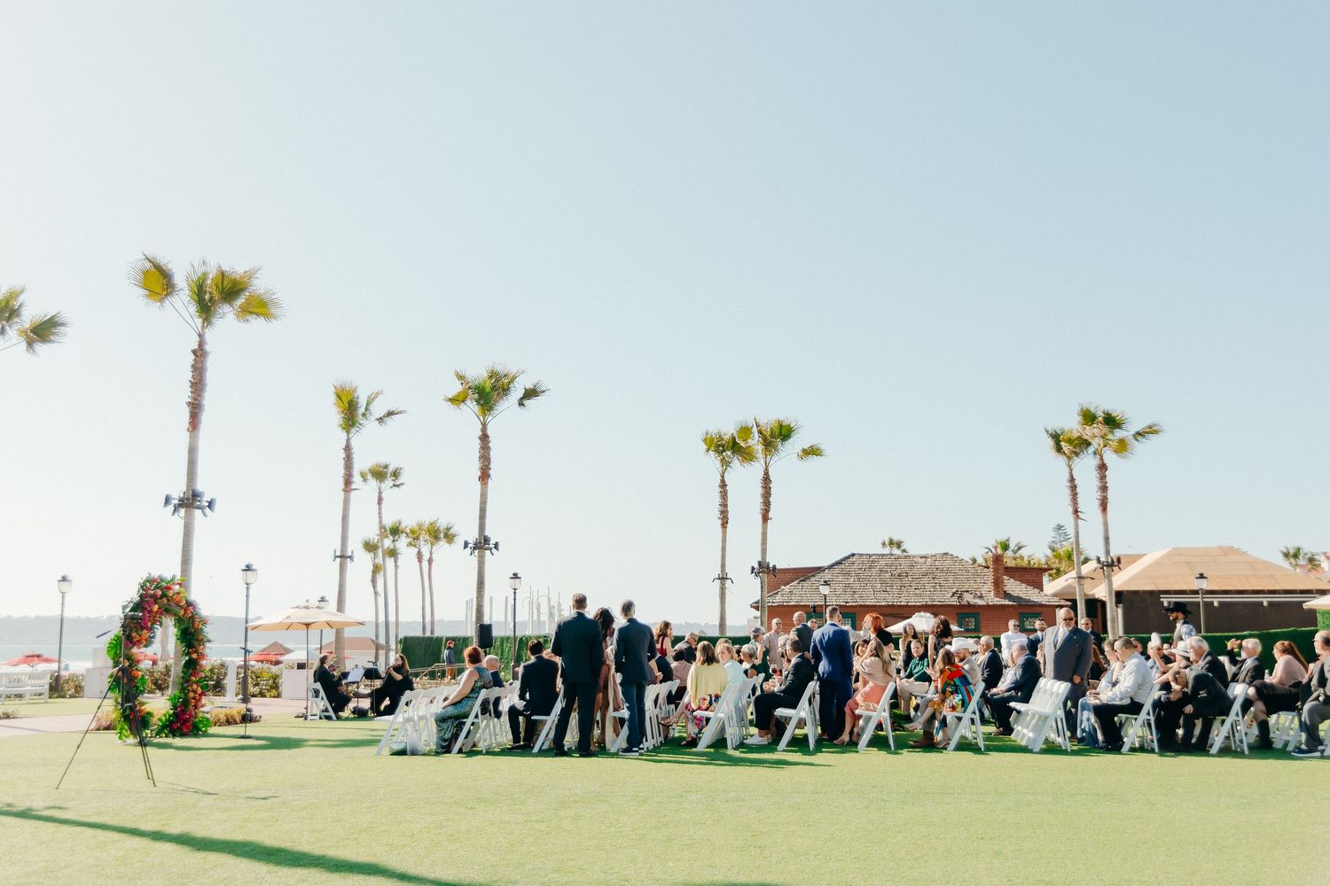 Wedding ceremony guests gather outdoors at a beachside venue with palm trees and ocean views on a sunny day.