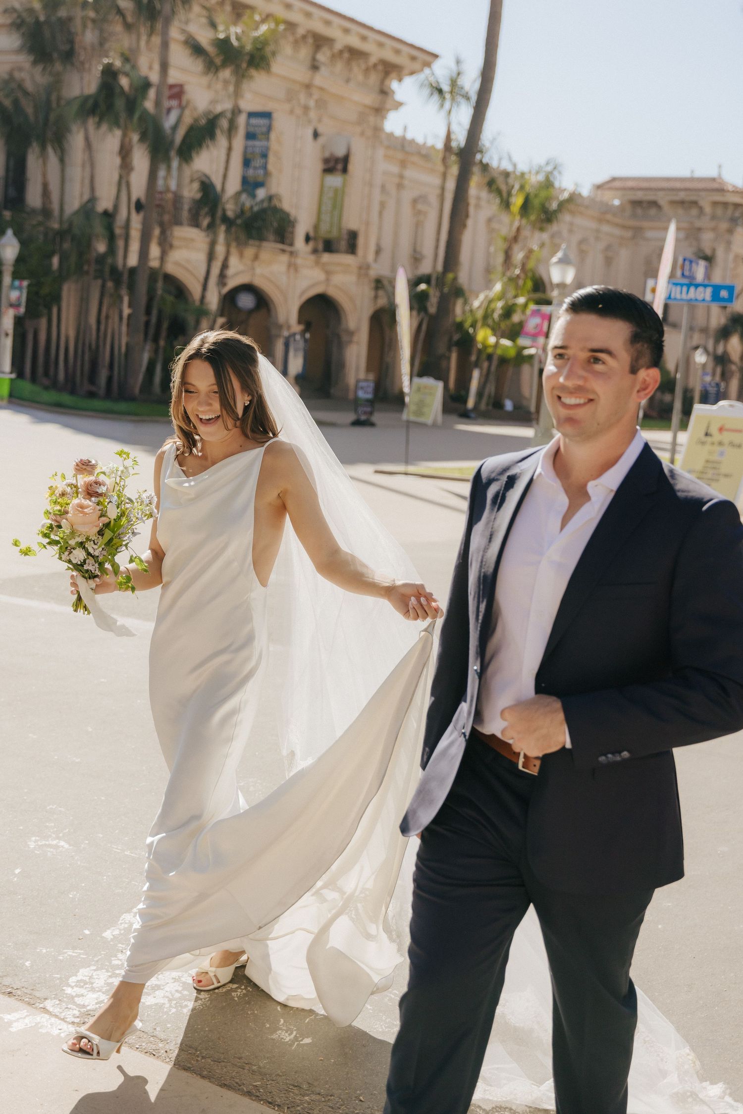 A couple in wedding attire walk together on a sunny day with palm trees and Spanish colonial architecture in the background.