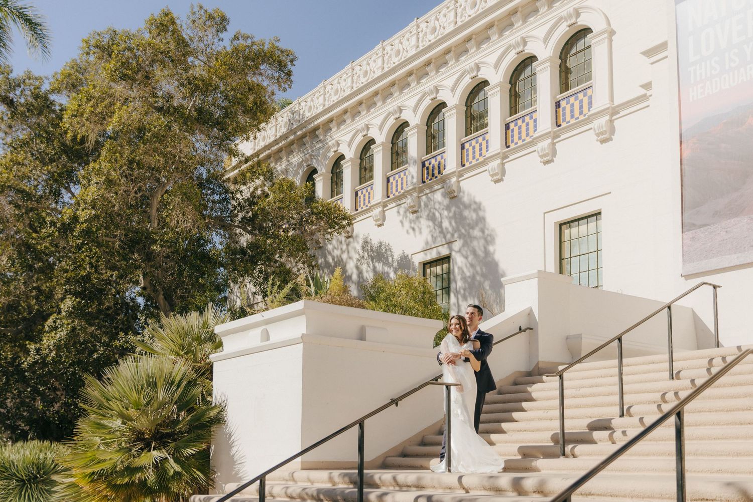 A couple embraces on the grand white stone staircase of an elegant Mediterranean-style building with arched architecture.