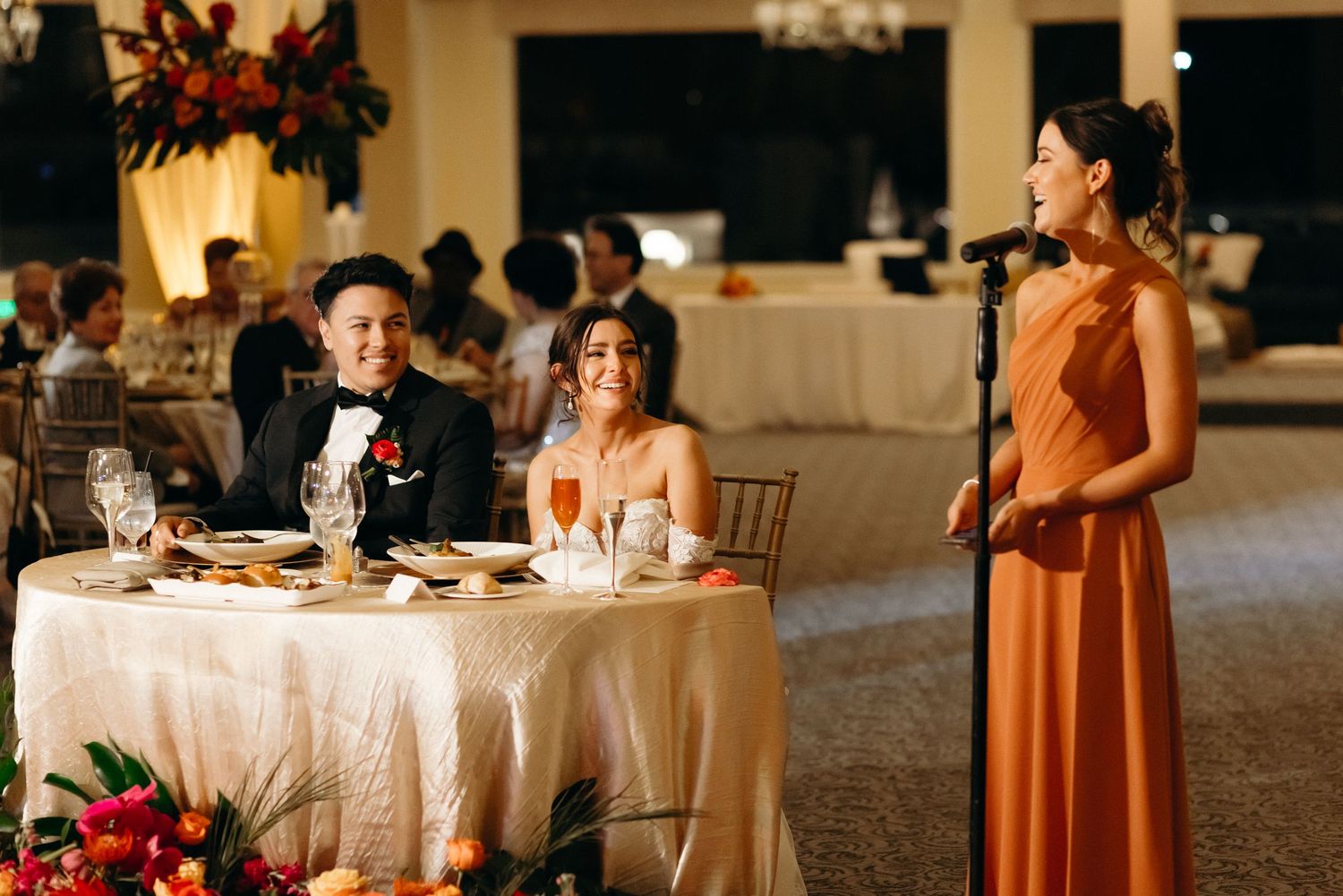 Reception scene with guests at table listening to speech in elegant ballroom setting.