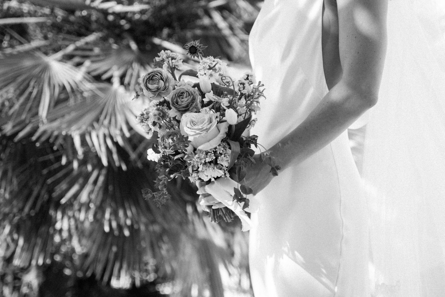 A couple in wedding attire shares a romantic moment in front of a sunlit garden with lush greenery.