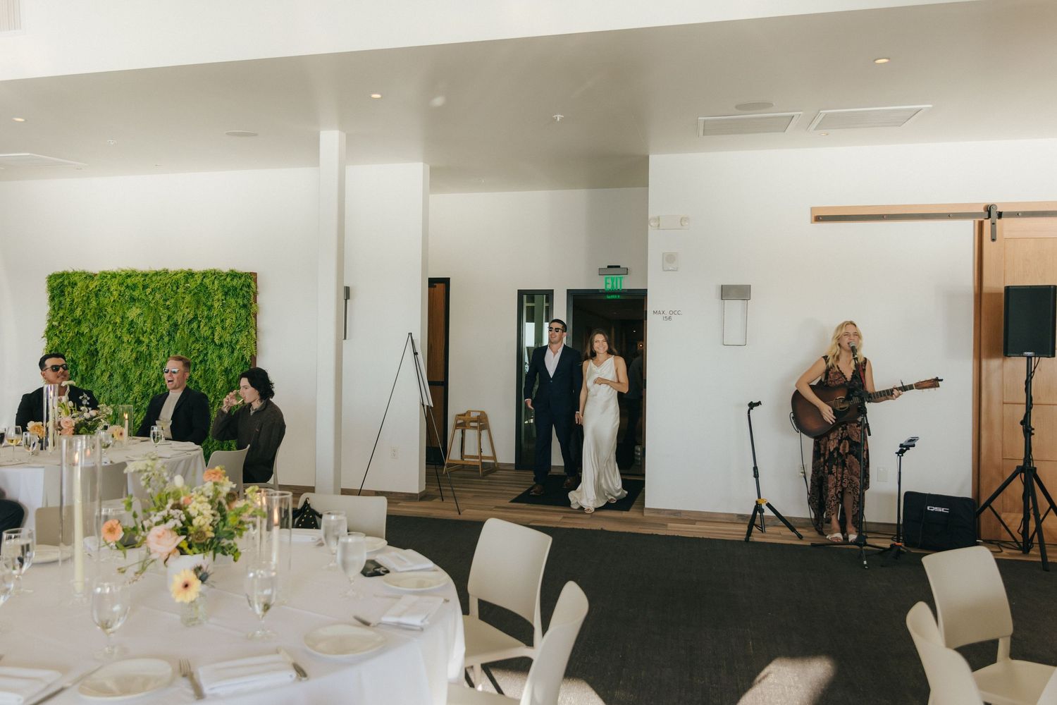A modern event space with white walls, dining tables, and a living plant wall backdrop during a celebration.