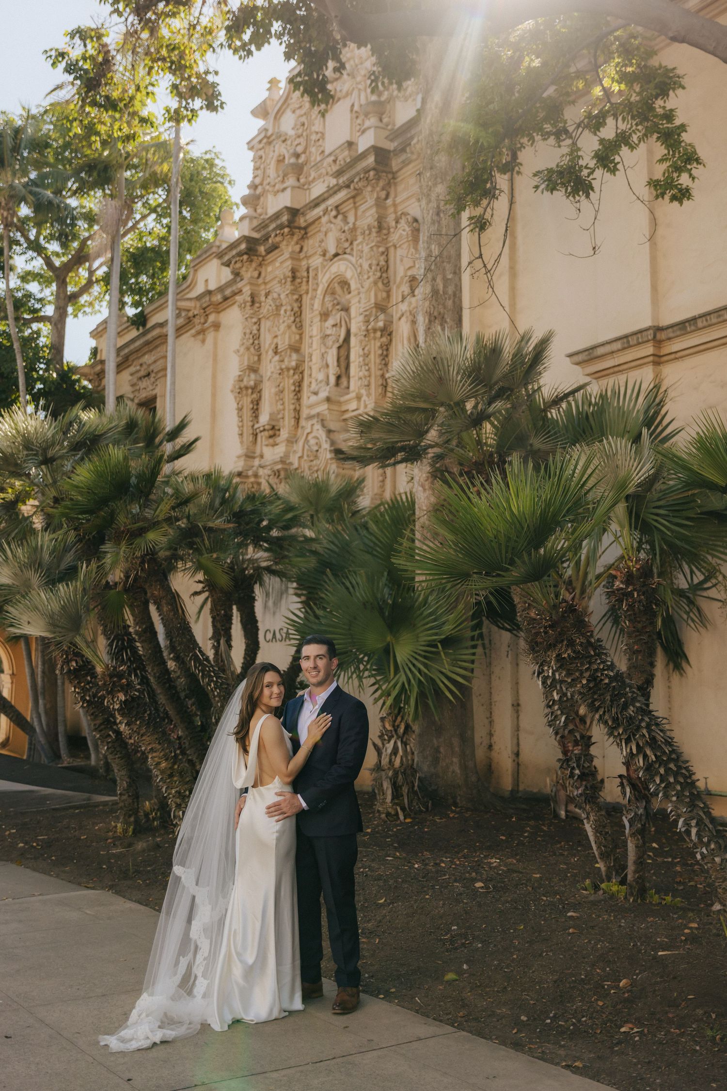A couple shares an intimate moment in front of a Spanish colonial building with palm trees and ornate architecture.