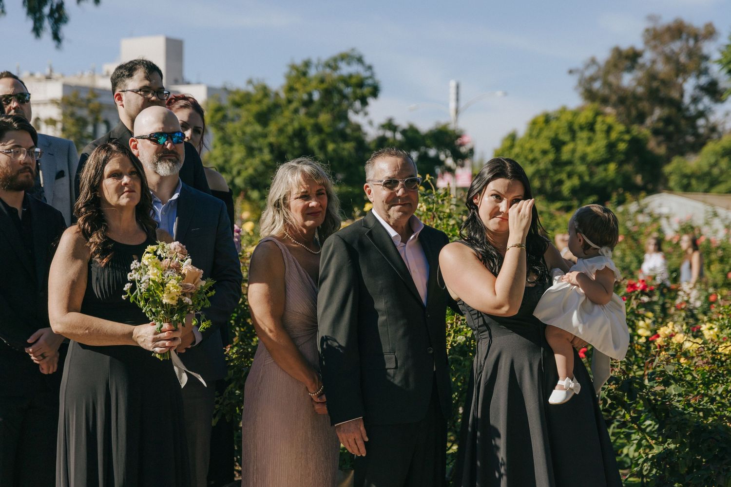 Wedding party stands together outdoors in formal attire with bridesmaids holding yellow bouquets in a garden setting.
