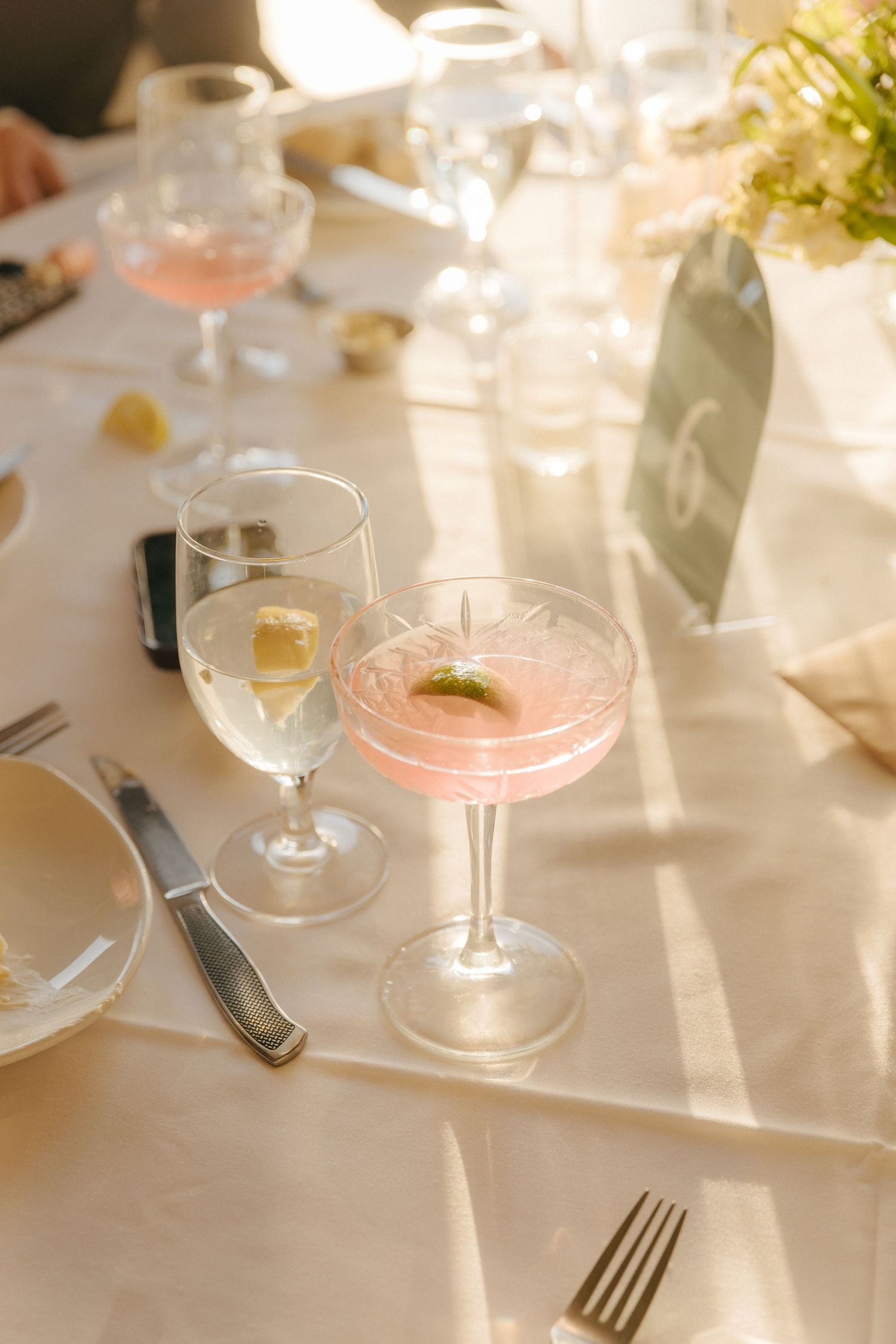 Elegant table settings with pink cocktails in coupe glasses and white wine glasses on a sunlit white tablecloth.
