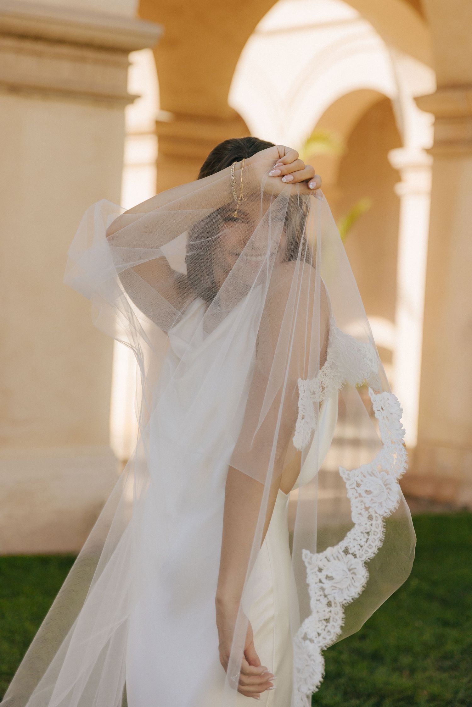 A flowing bridal veil with delicate lace detail catches sunlight beneath arched colonnade walkways.