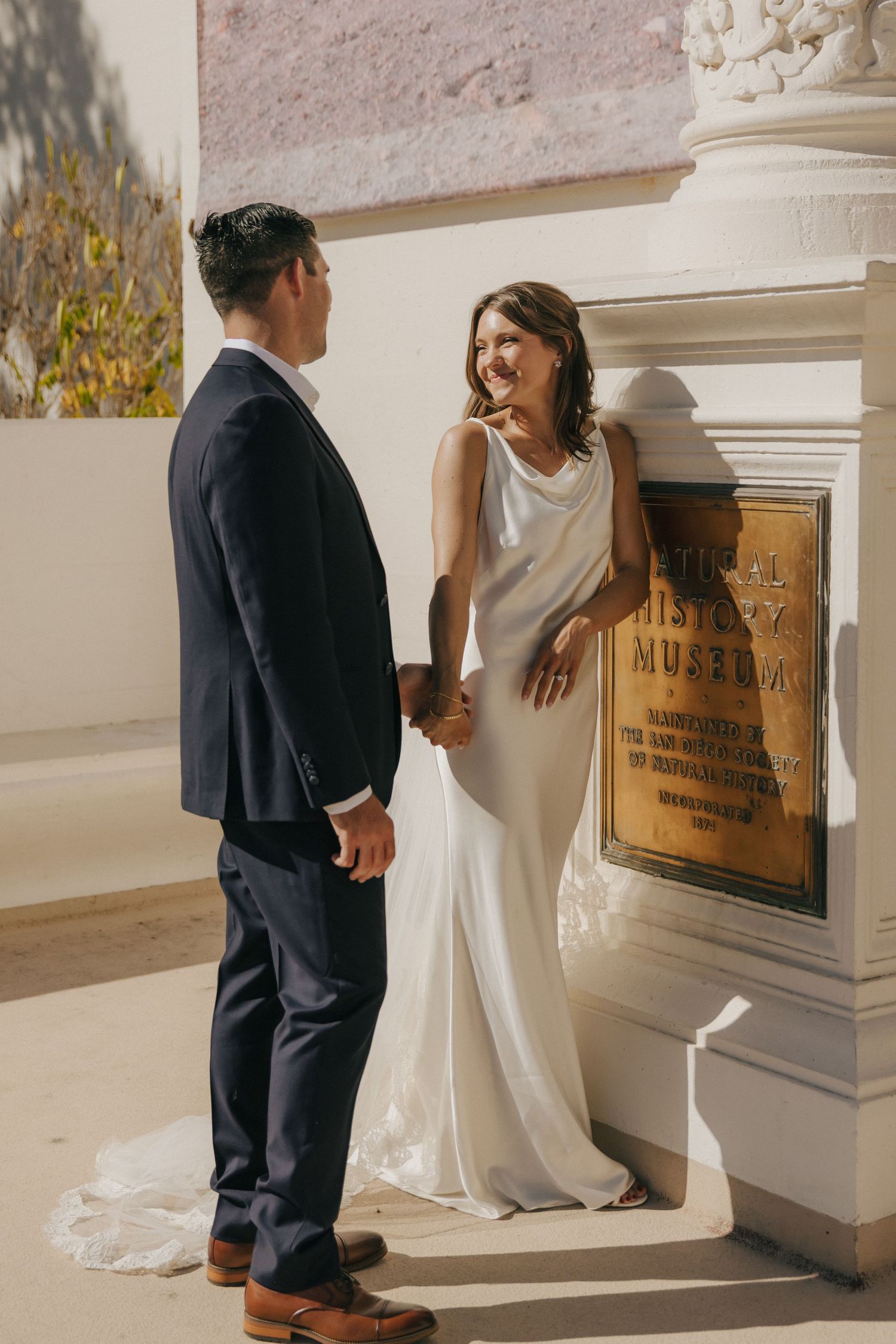 A bride in a white dress and groom in a dark suit share a tender moment near an ornate architectural column.