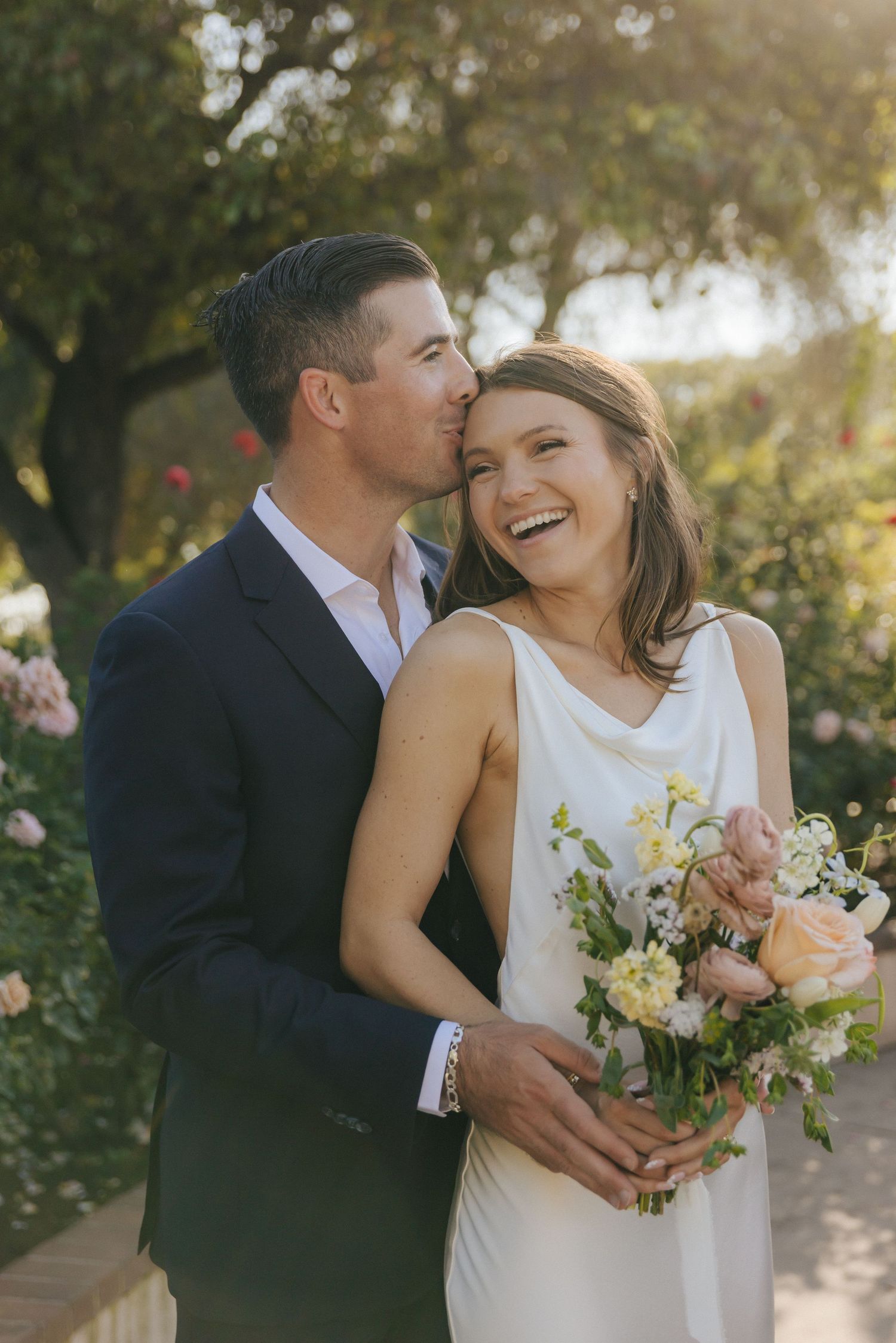 A romantic wedding couple embraces outdoors in natural lighting with the bride holding a delicate white floral bouquet.
