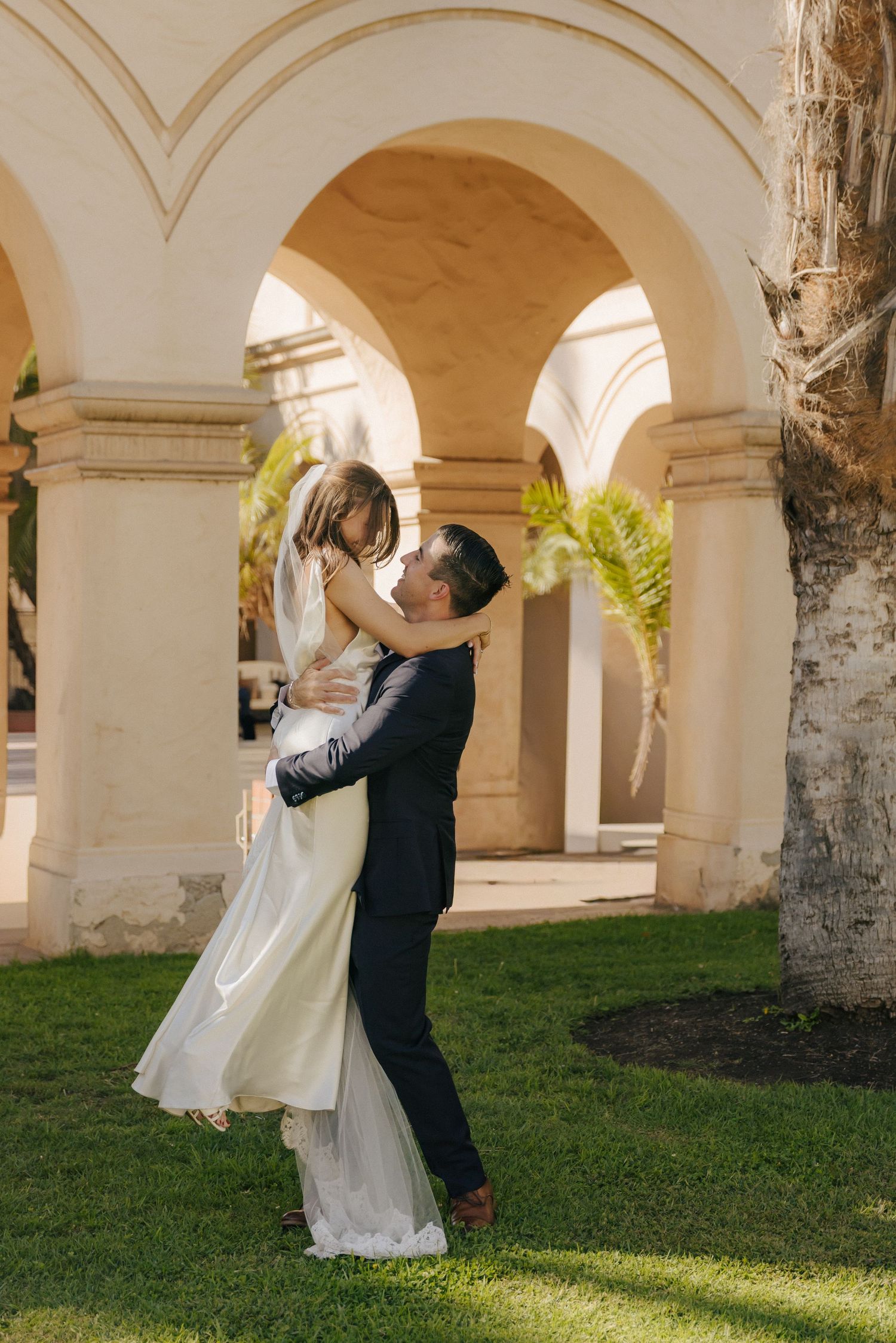 A couple dances together under arched columns and palm trees in golden sunlight.