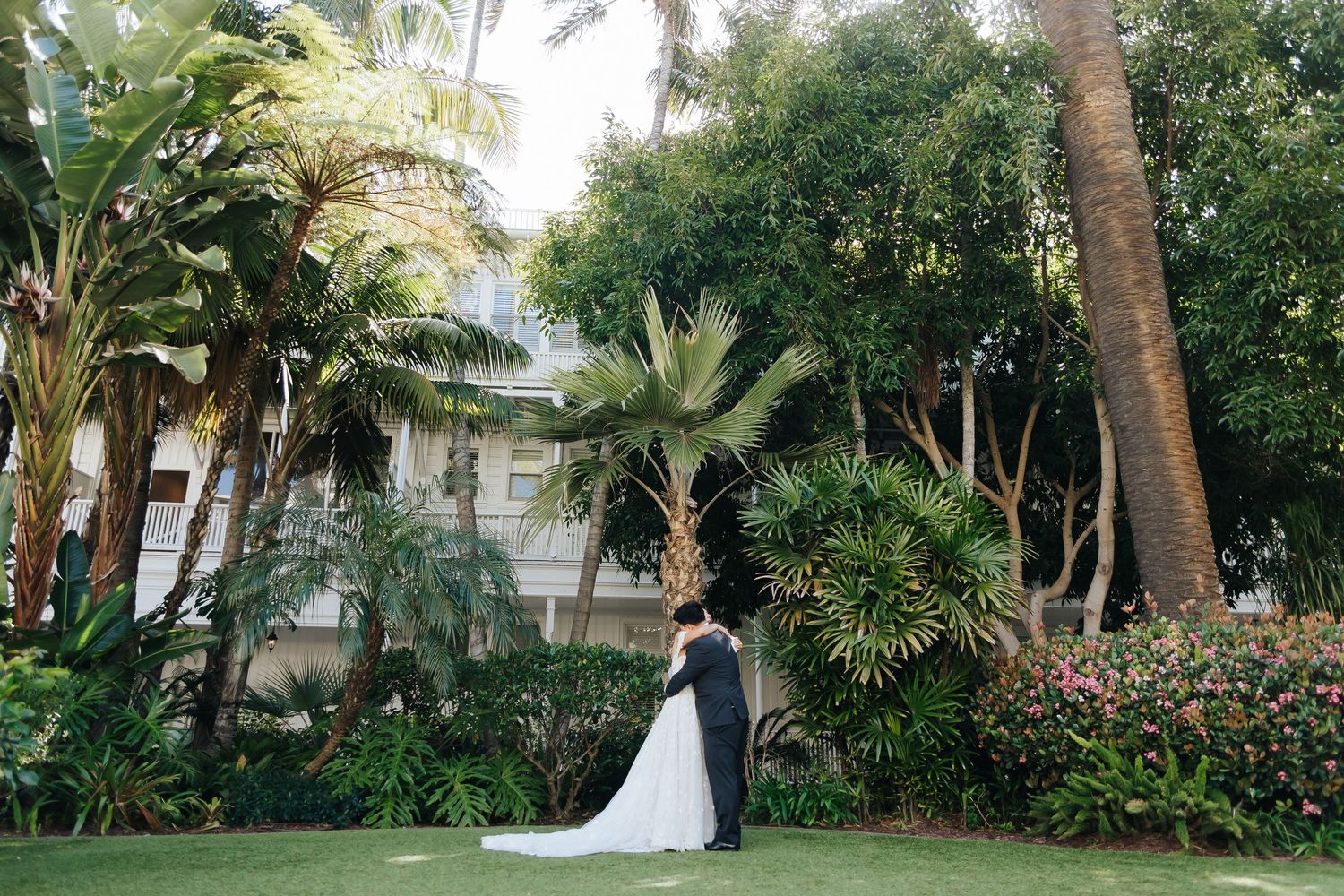 A couple embraces in front of a tropical garden with palm trees and lush greenery outside a white house.