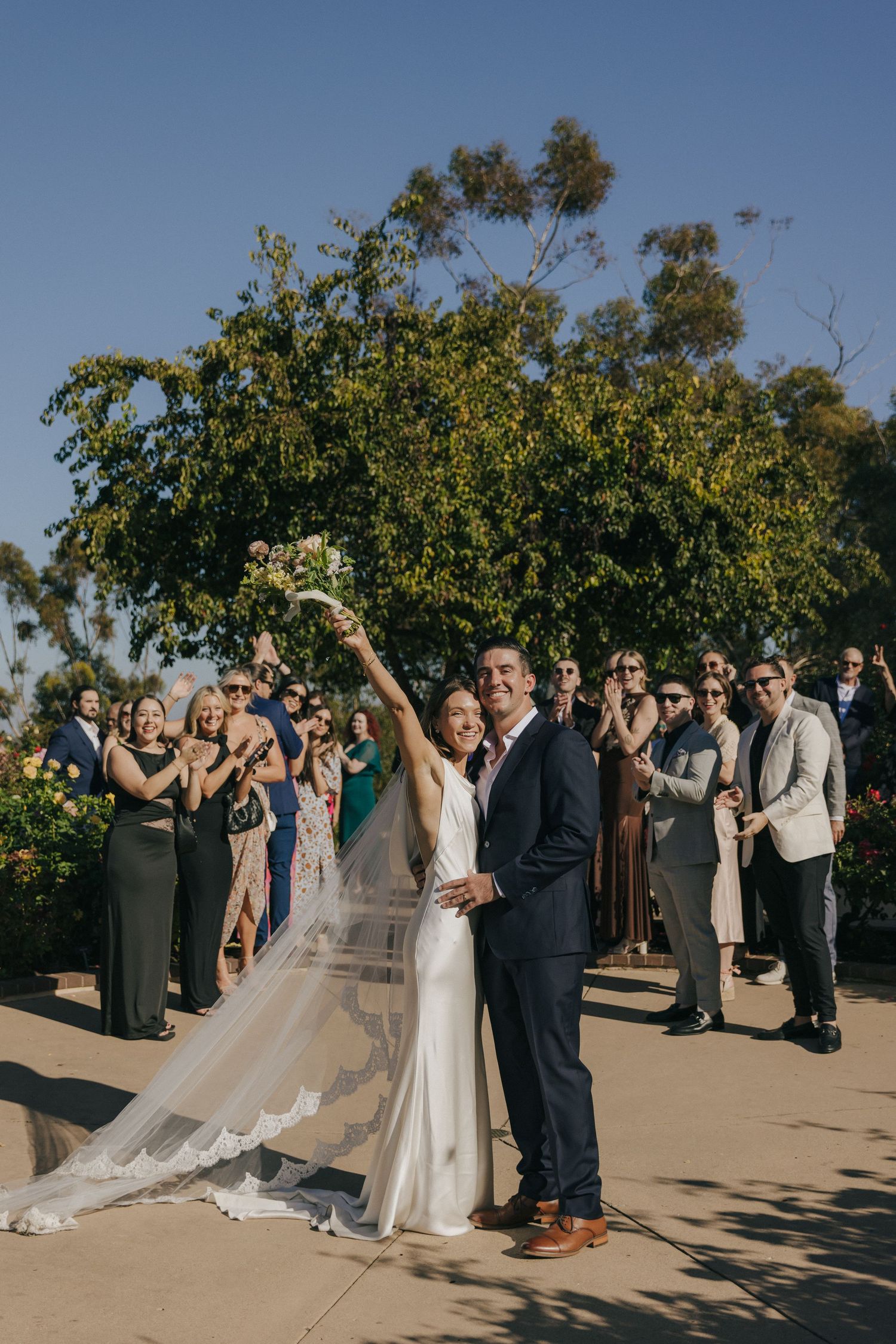Wedding ceremony taking place outdoors on sunny day with guests gathered under tree and long flowing veil.