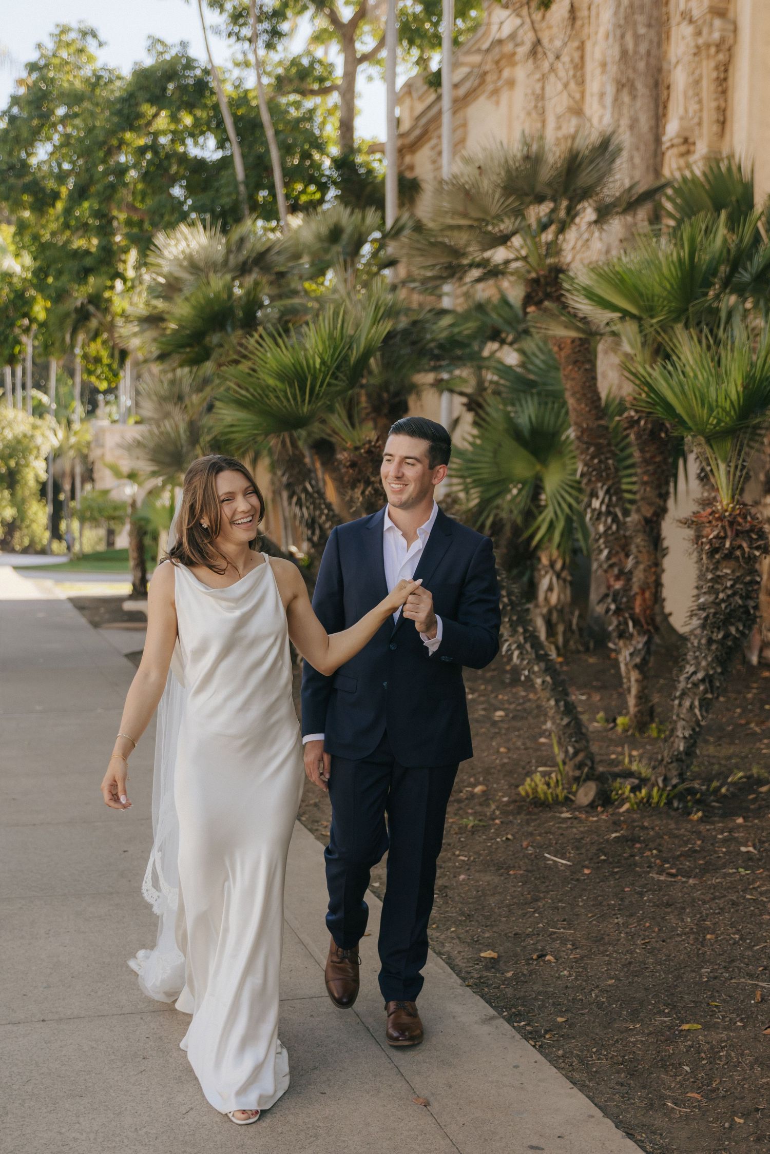 A couple playfully walks together along a palm tree-lined path during their wedding photoshoot.