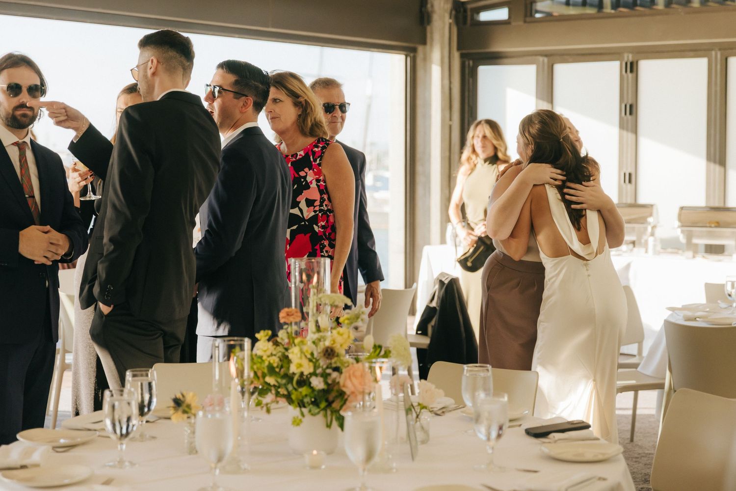 Wedding guests mingle around an elegantly set dining table with floral centerpiece in a bright sunlit venue.