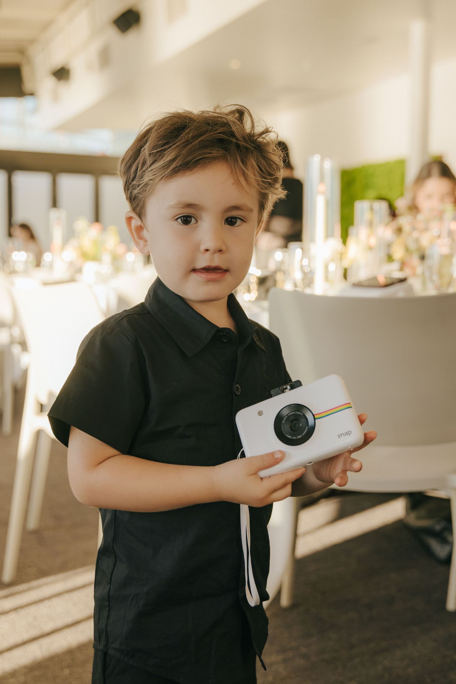 A small child in black clothing holds a white camera at an indoor event.