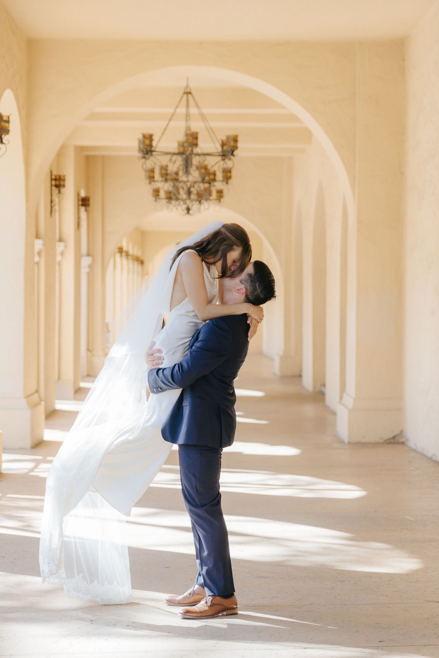 Wedding couple shares intimate moments in a sun-lit colonnade with white arches and chandeliers.