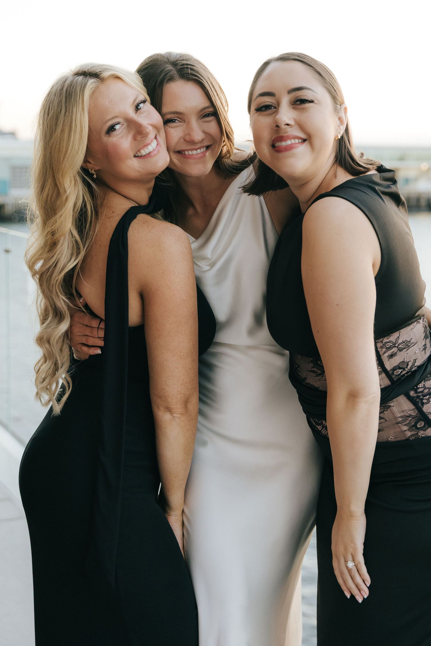 A group of friends in black and white dresses laugh and embrace while posing together for photos outdoors.