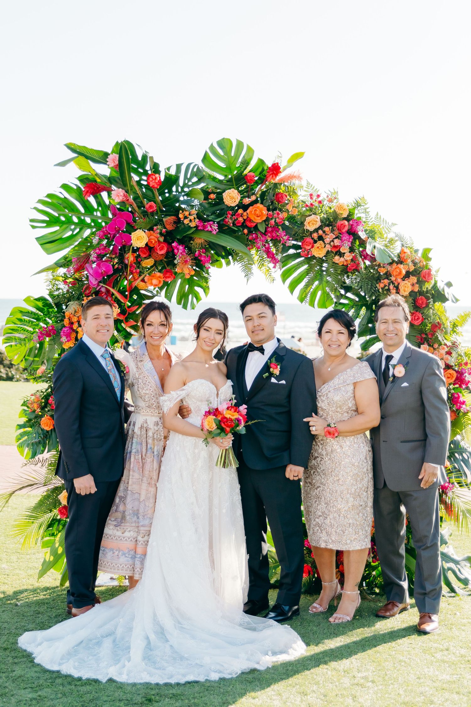 Wedding party poses under vibrant tropical floral arch with monstera leaves and colorful blooms.