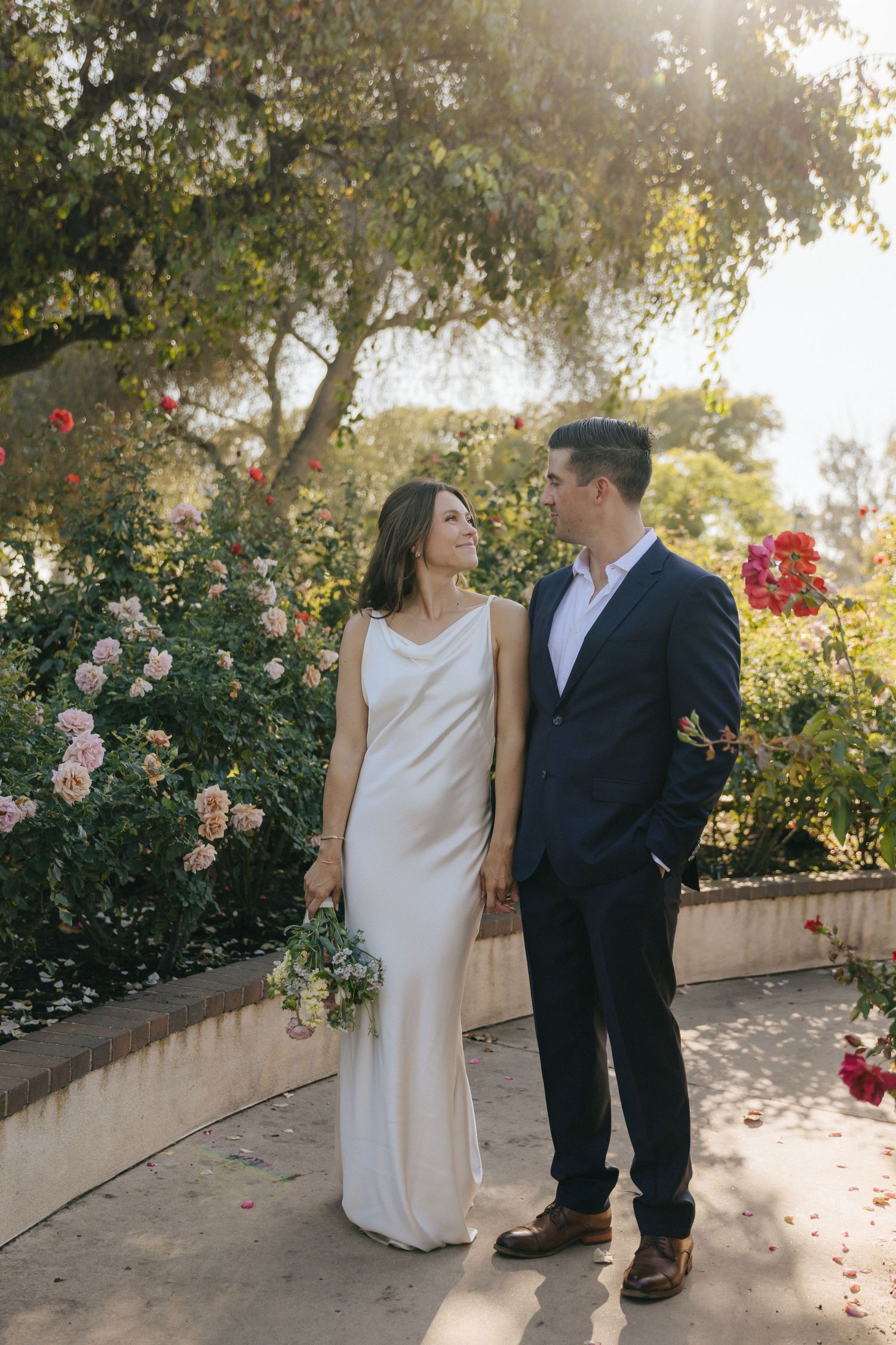 A bride in white and groom in navy share tender moments against a backdrop of blooming roses.