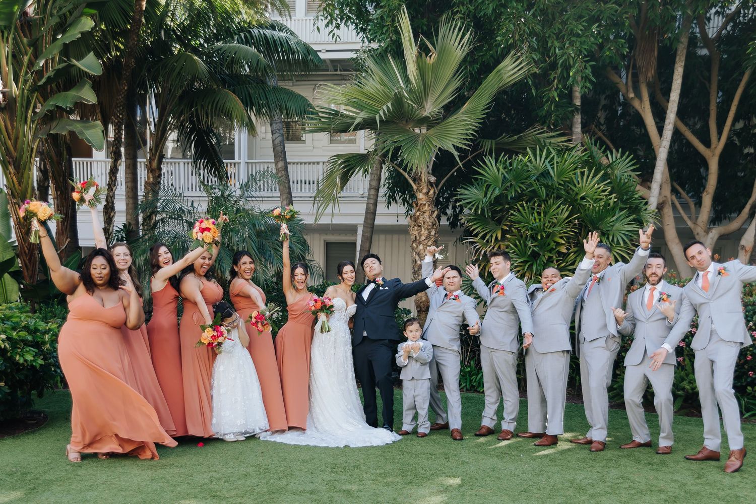 Wedding party in coral bridesmaids dresses and light grey suits pose together on a tropical garden lawn.