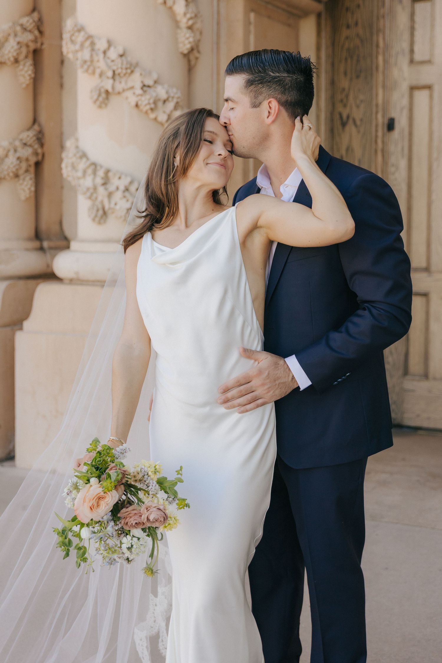Romantic wedding couple shares intimate moments in an elegant colonnade while holding delicate floral bouquets.