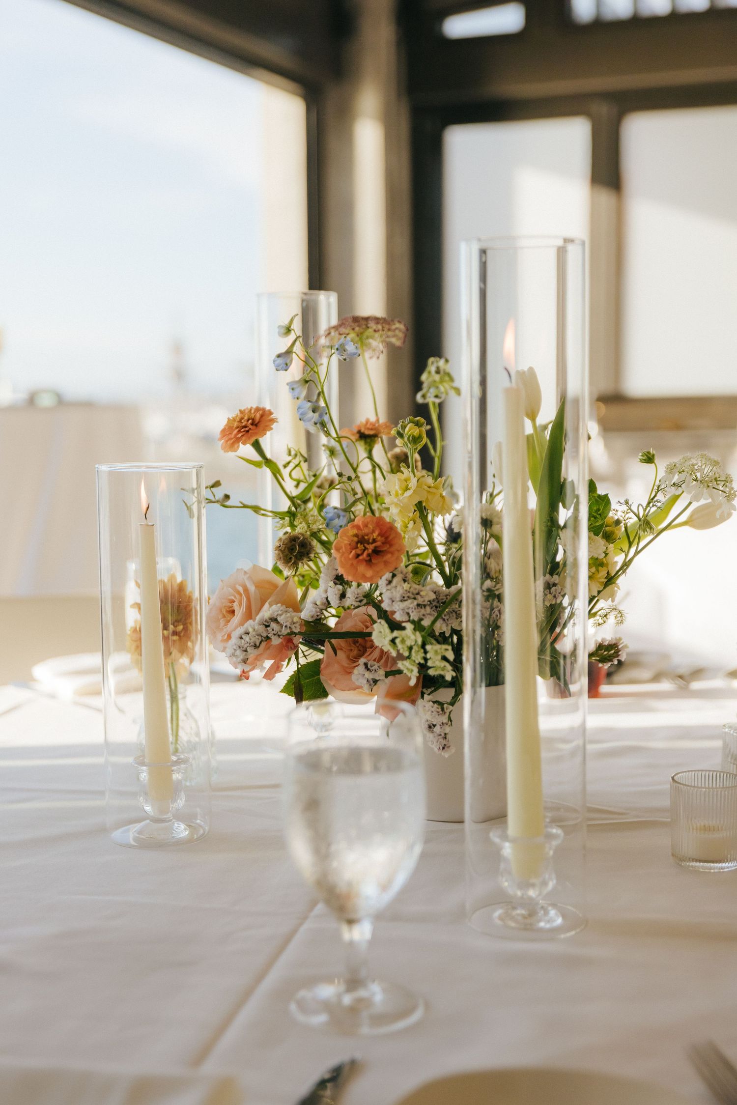 Elegant table setting with tall white candles and peach floral centerpiece arrangements on crisp white linens.