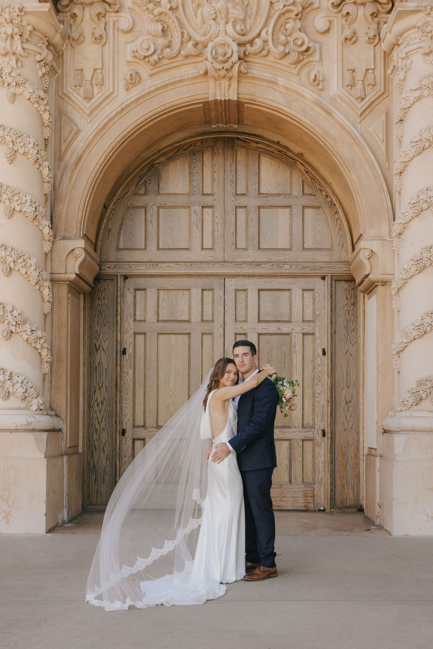 Romantic wedding couple embraces under ornate stone archways in a historic architectural setting.
