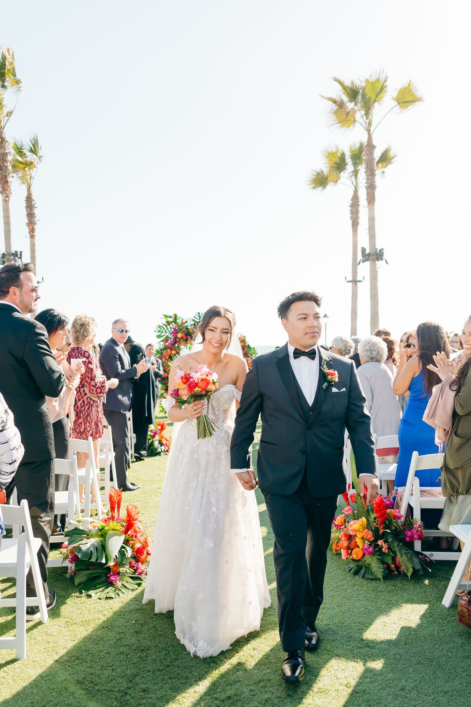 Newlyweds walking down the aisle surrounded by guests and tropical floral arrangements.