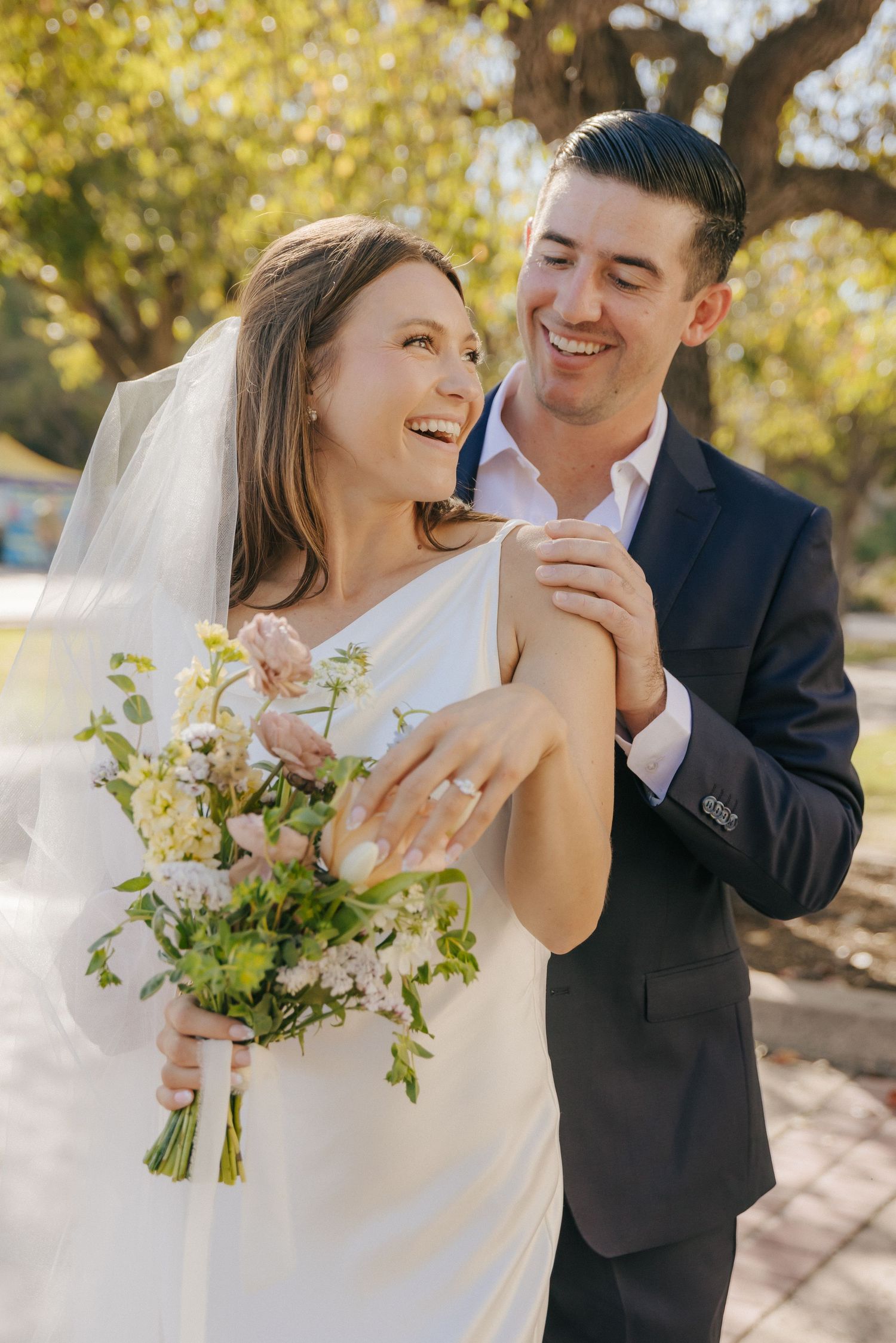 Joyful wedding couple laughs together in warm sunlight while holding a spring bouquet.