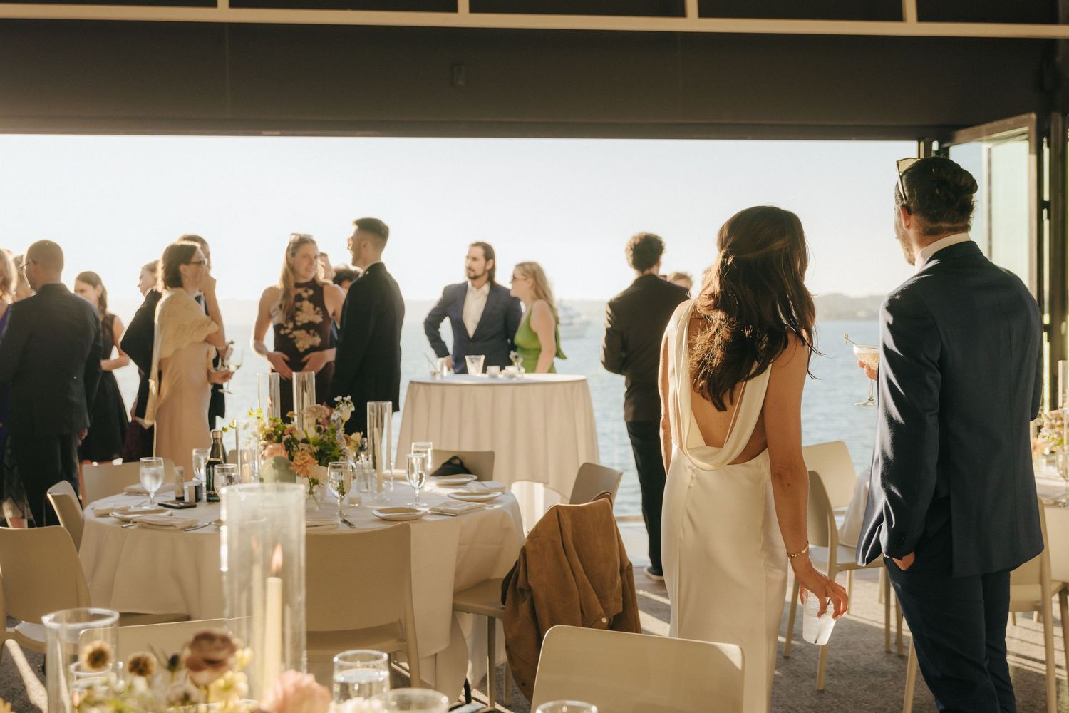 Wedding guests mingle and socialize at an elegant reception with white tables and chairs in a bright venue.