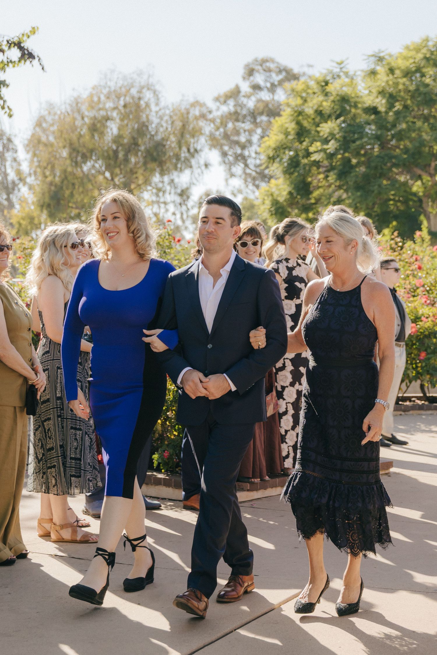 A group of wedding guests walk together on a sunny patio, dressed in formal attire including blue and black dresses.