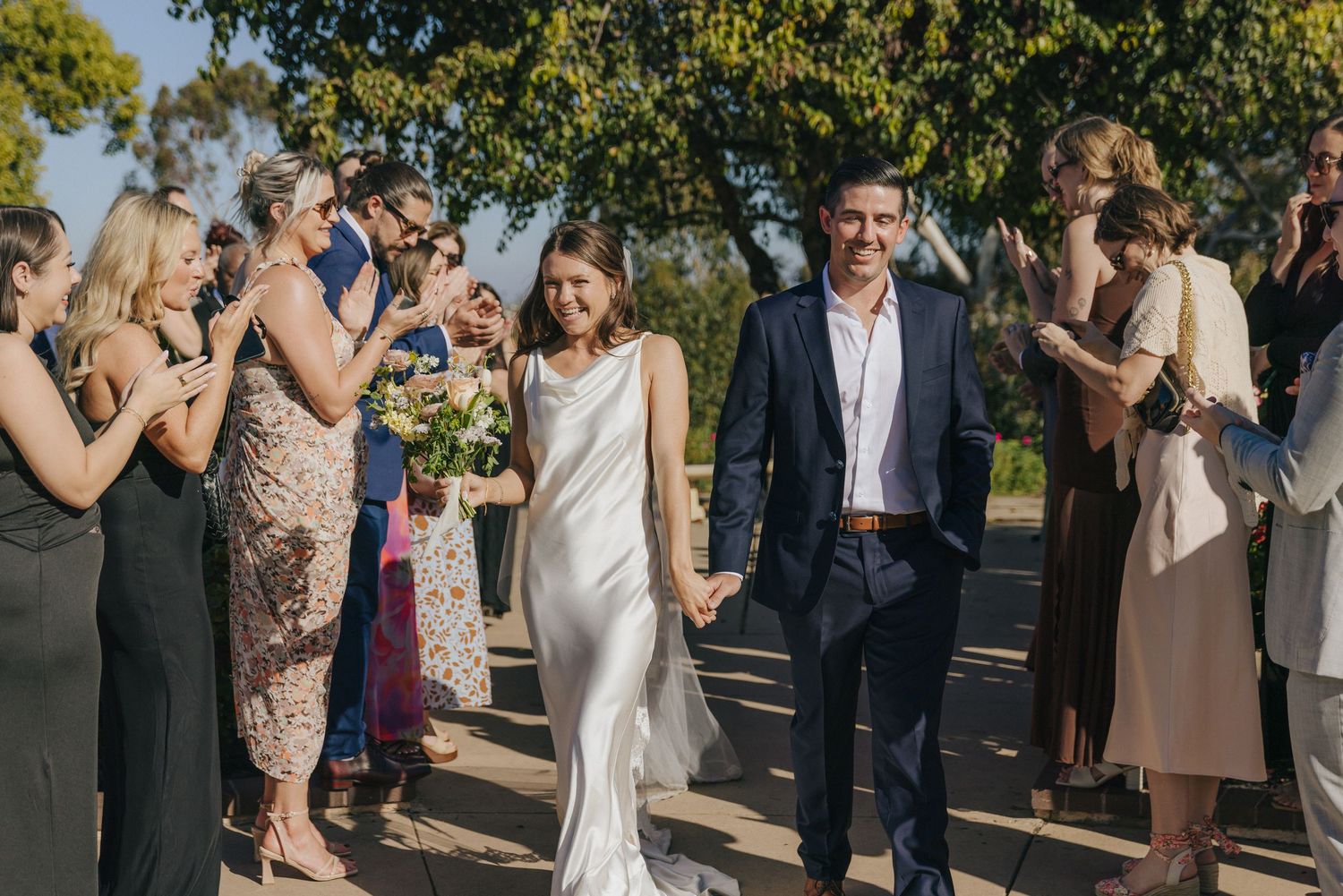 Wedding guests in formal attire applaud as a couple walks down an outdoor pathway on a sunny day.