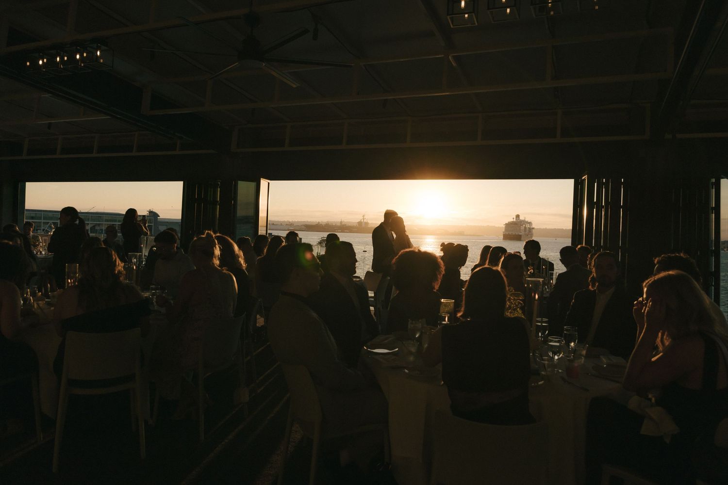 A silhouetted crowd gathers at an indoor venue during sunset, creating a warm atmospheric scene through large windows.