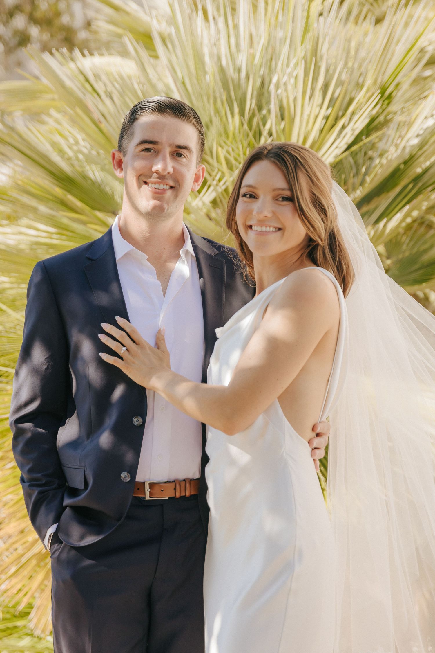 A wedding couple poses outdoors in front of palm fronds, with the bride in white and groom in navy suit.