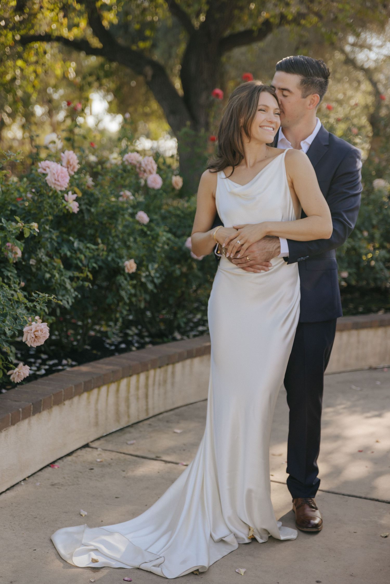 A couple embraces in a garden setting with pink roses, the bride wearing a flowing white gown and groom in navy suit.