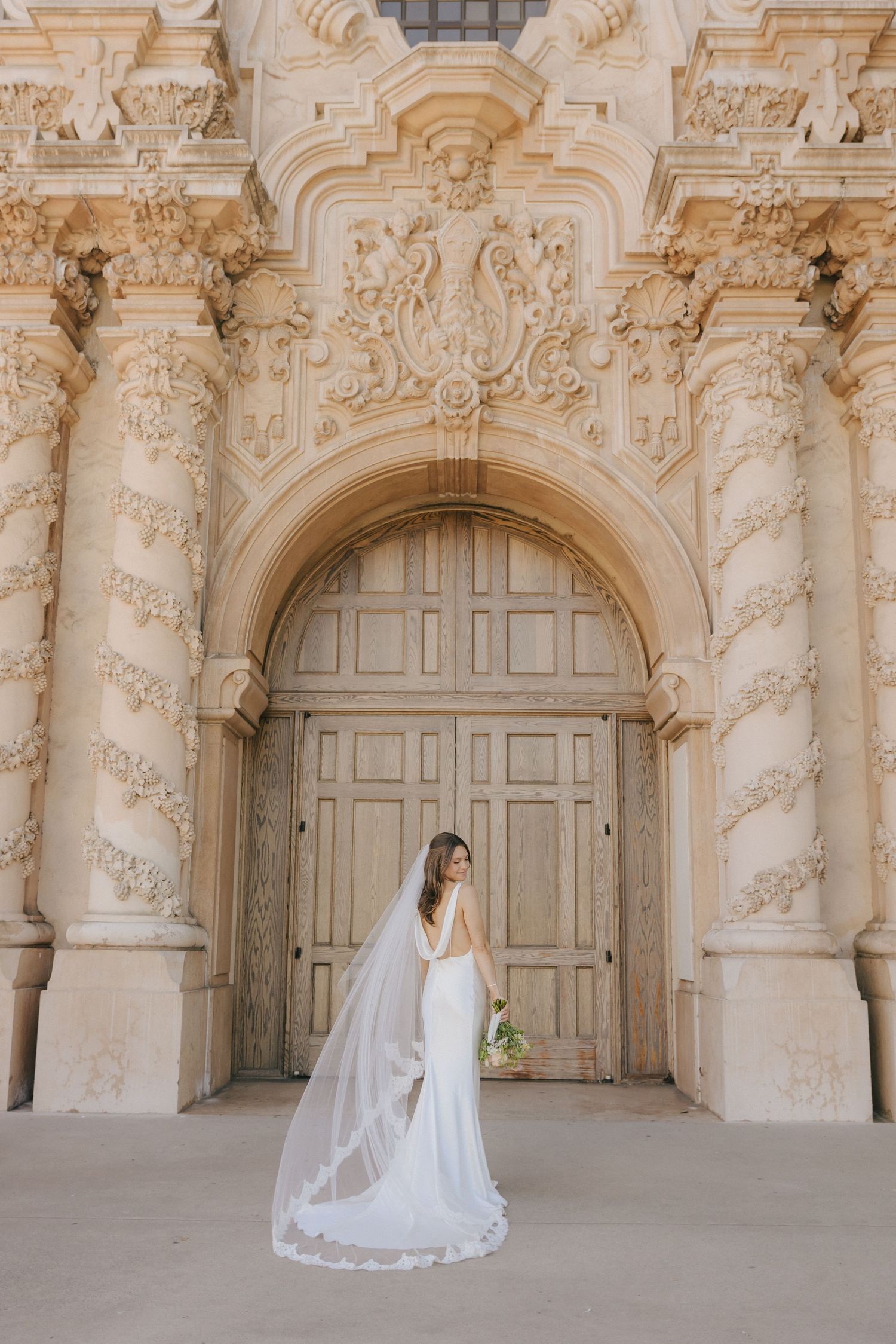 A bride in a flowing white gown and veil poses beneath ornate baroque archways at a historic venue.