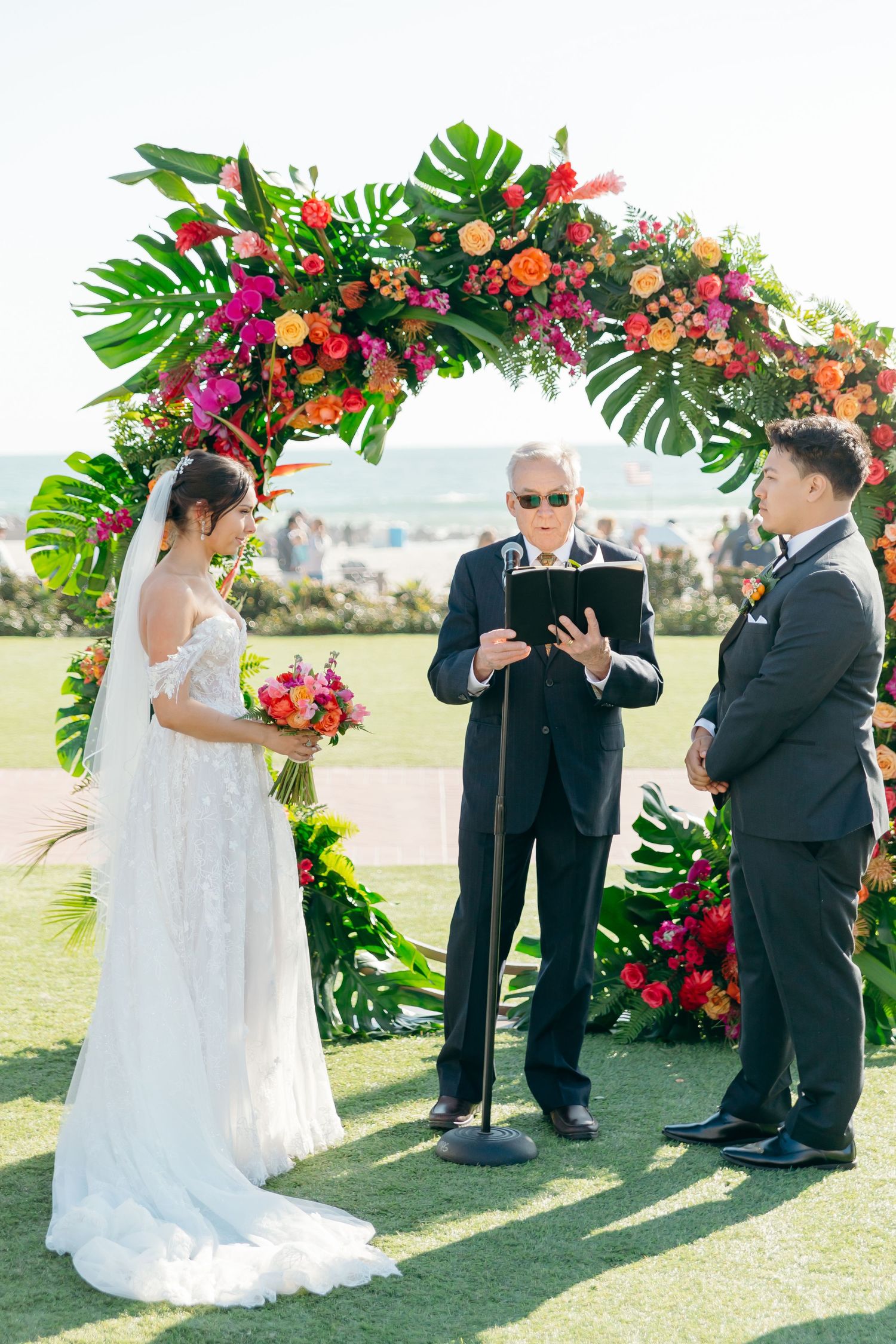A wedding ceremony under a tropical floral arch with monstera leaves and colorful flowers on a grassy outdoor venue.