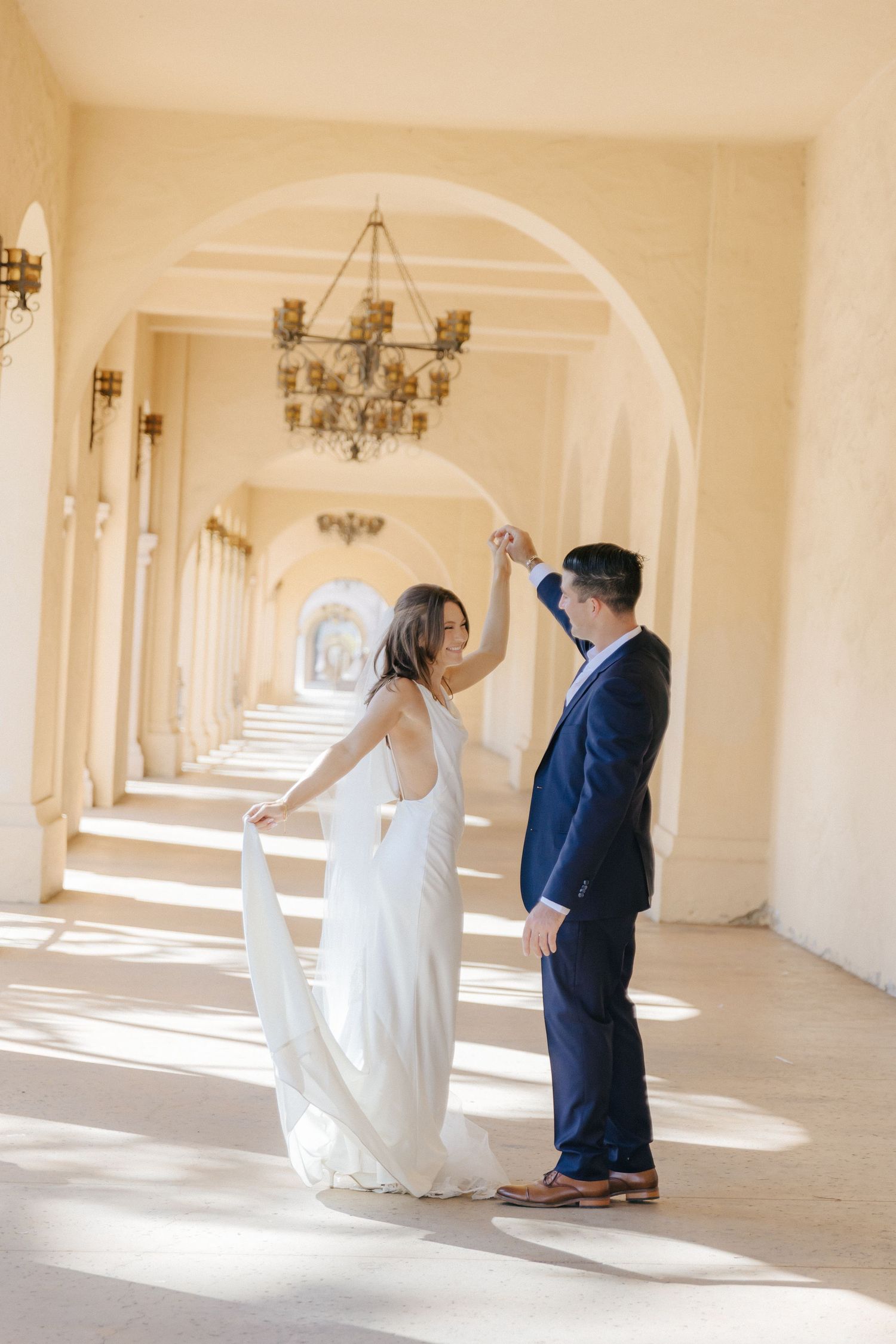 A couple dancing in an elegant hallway with arched columns and chandeliers as sunlight streams through the windows.