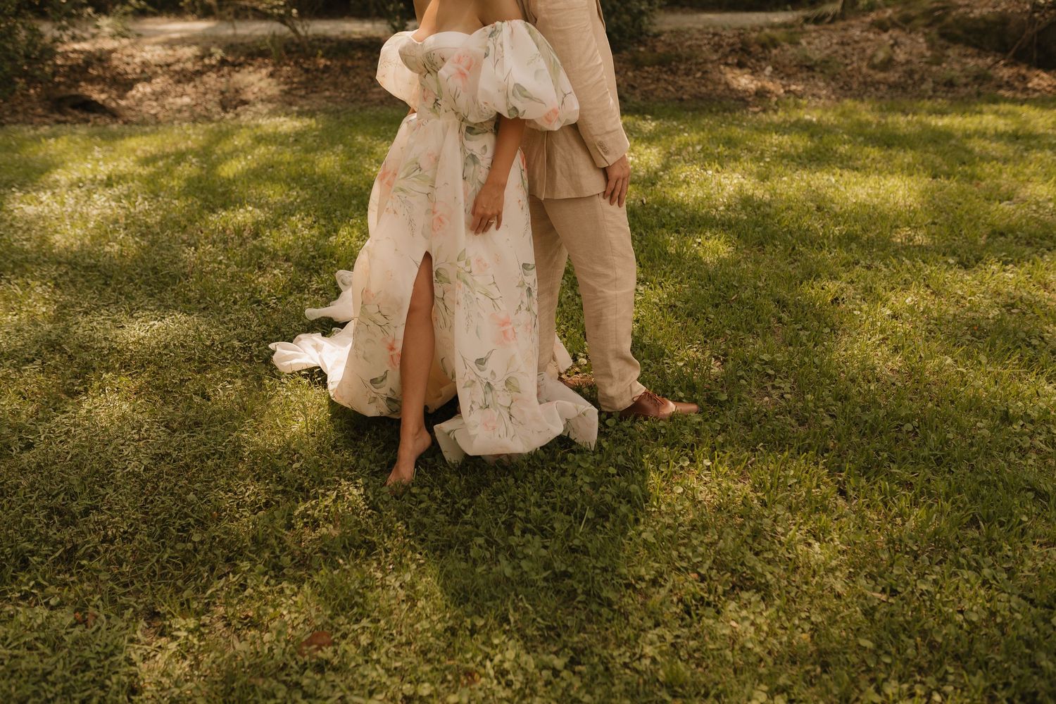 Barefoot couple in floral wedding dress and tan pants standing on lush green grass.