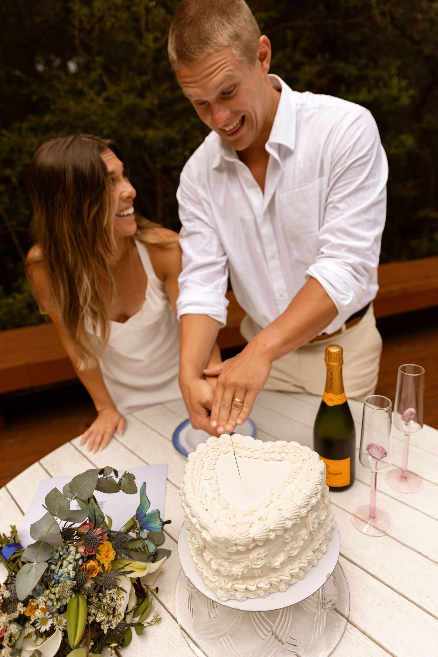 Smiling couple cutting a heart-shaped white wedding cake outdoors, with champagne and floral bouquet on a white table.