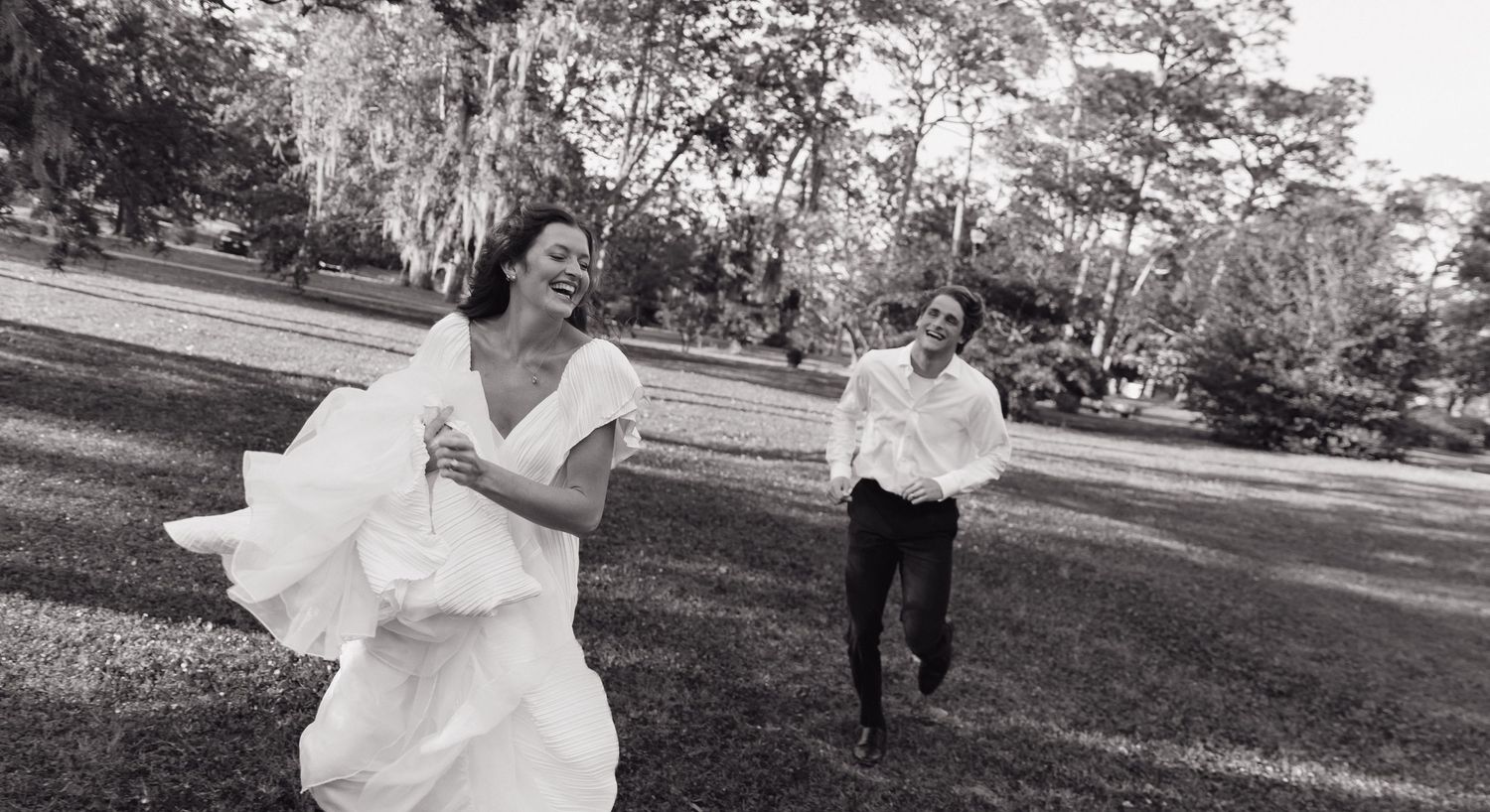Joyful bride in white gown runs laughing with groom through green park in black and white photo.