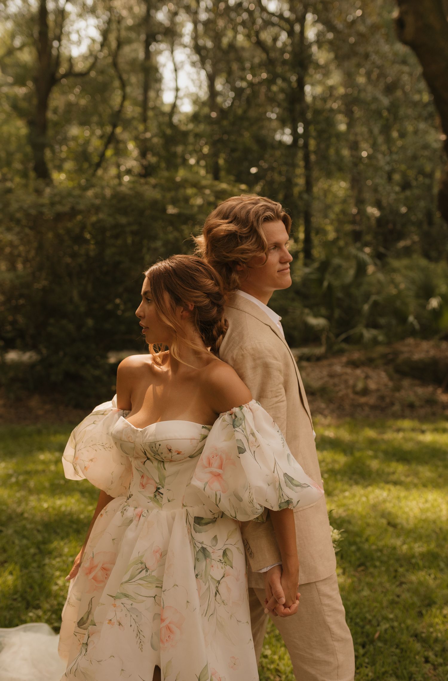A bride in a floral off-shoulder gown and groom in tan suit stand back-to-back in a lush garden setting.