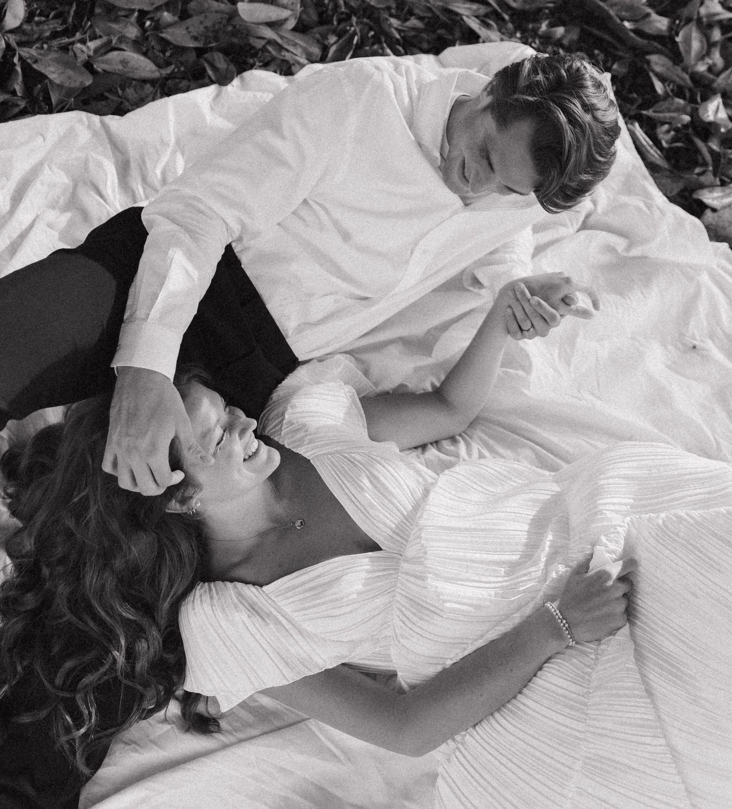 Black and white romantic photo of bride and groom lying together on fallen leaves outdoors.