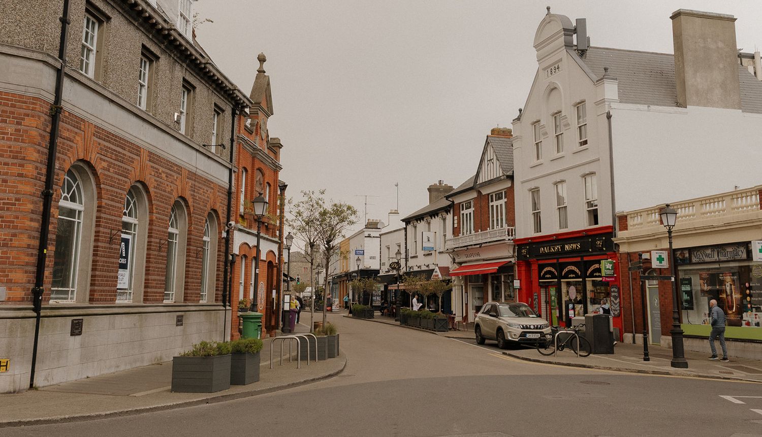 Charming town high street with red brick buildings, parked cars, and shops on a cloudy day.