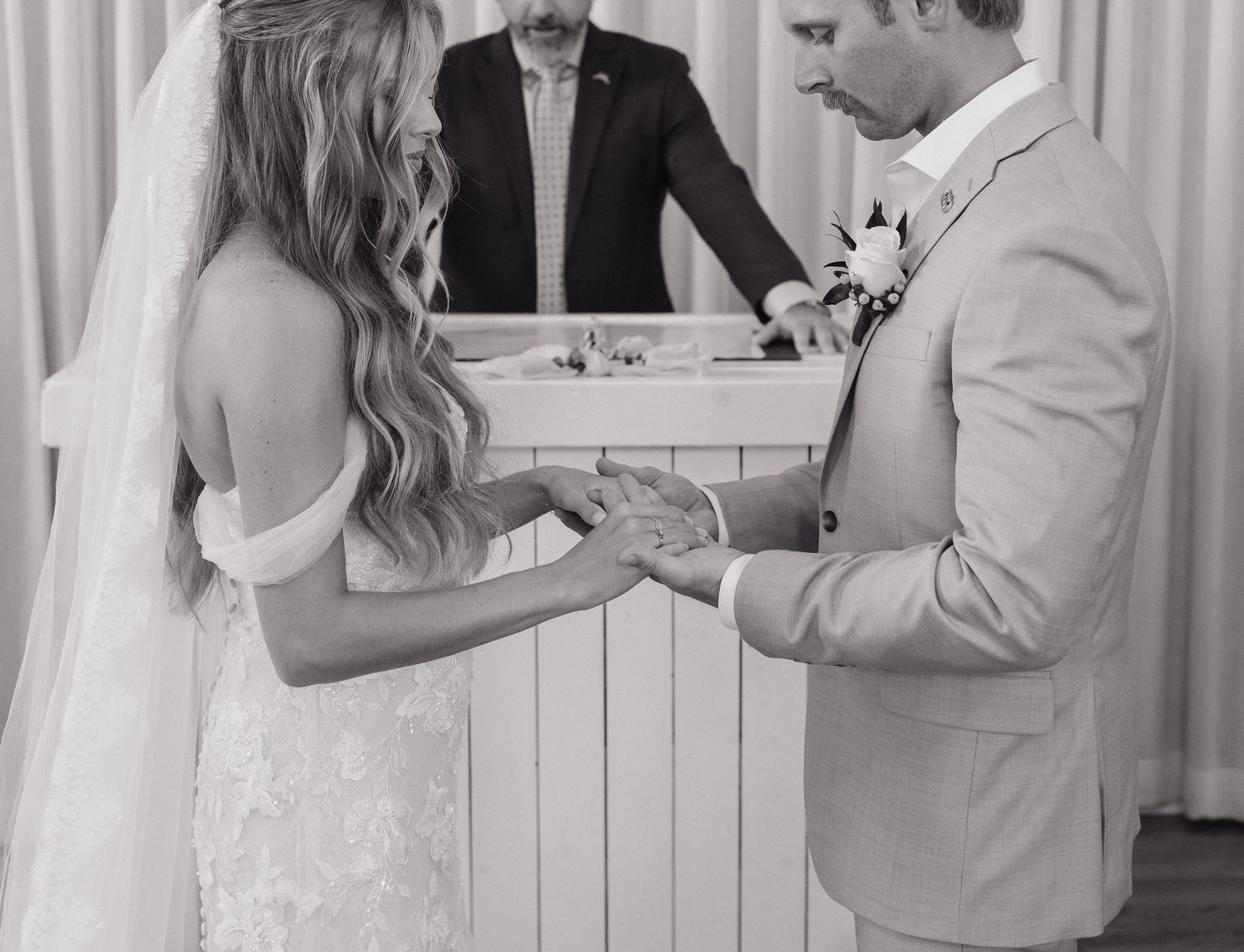 Black and white photo of bride and groom exchanging rings during wedding ceremony with officiant.