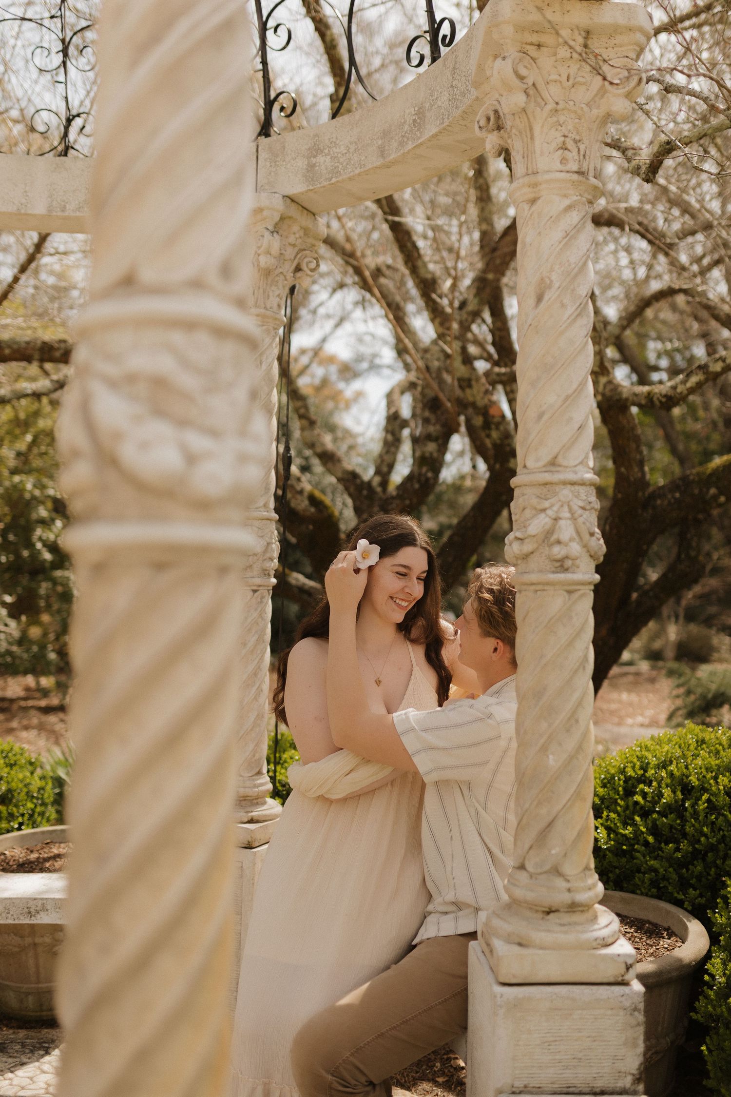 Couple sharing a romantic moment under ornate white stone columns in a garden setting.