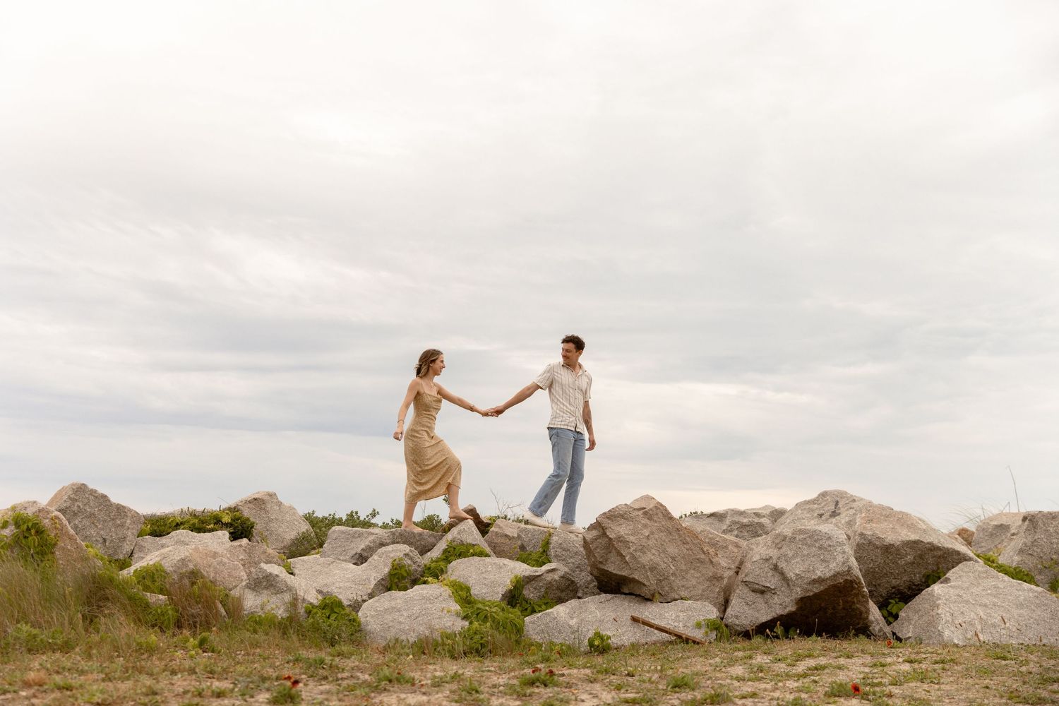 A couple holds hands while walking on rocky terrain under a cloudy sky in a romantic outdoor setting.