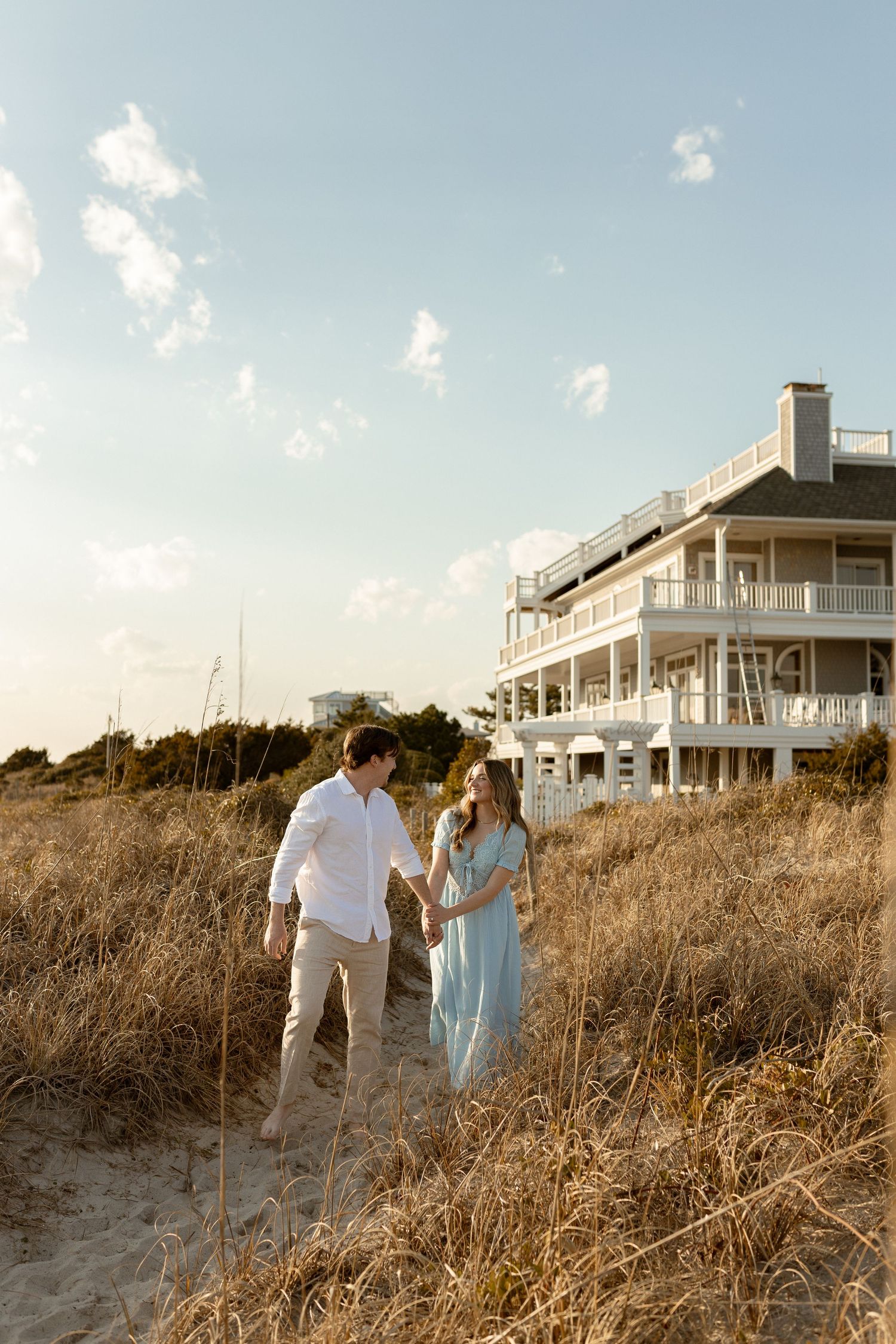 Couple holding hands walking through beach grass dunes with a large white beachfront house in the background.