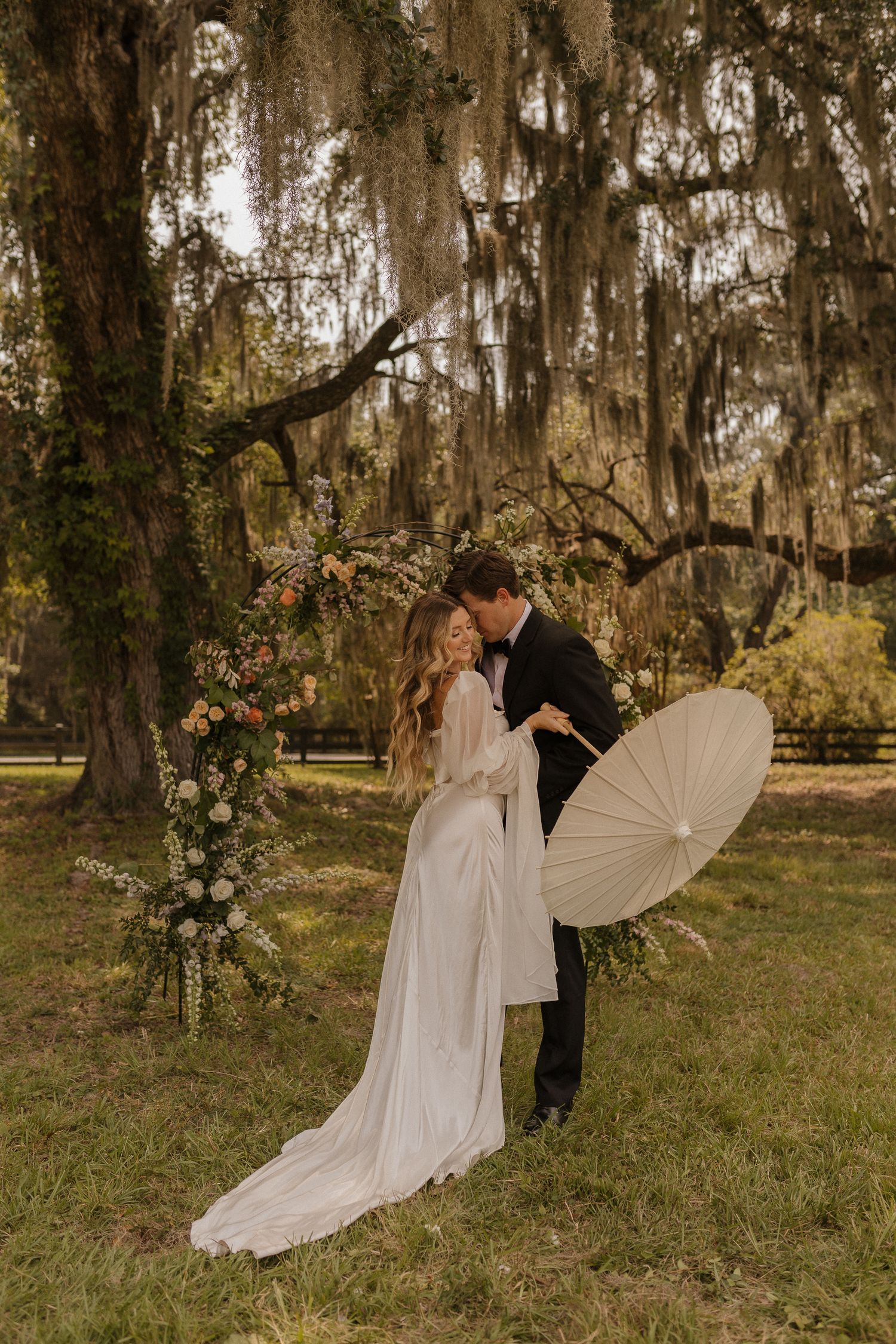 Bride and groom share a romantic moment under Spanish moss trees with a floral arch and parasol at an outdoor wedding.