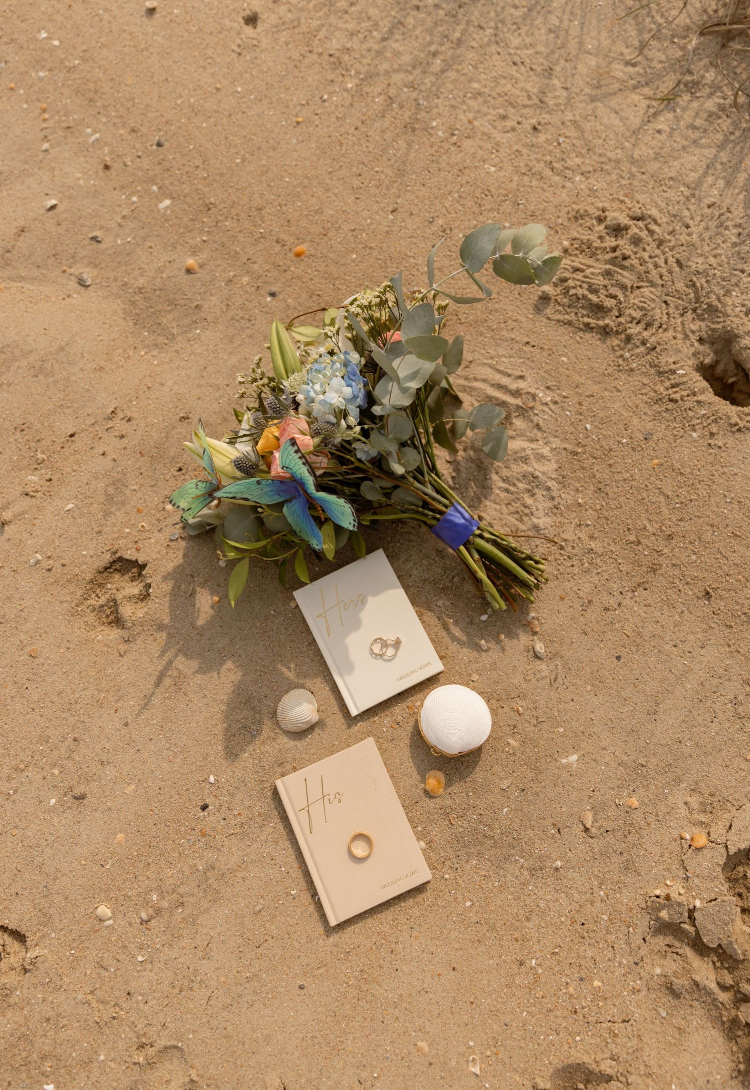 Wedding bouquet with blue flowers and eucalyptus beside rings and cards on sandy beach.